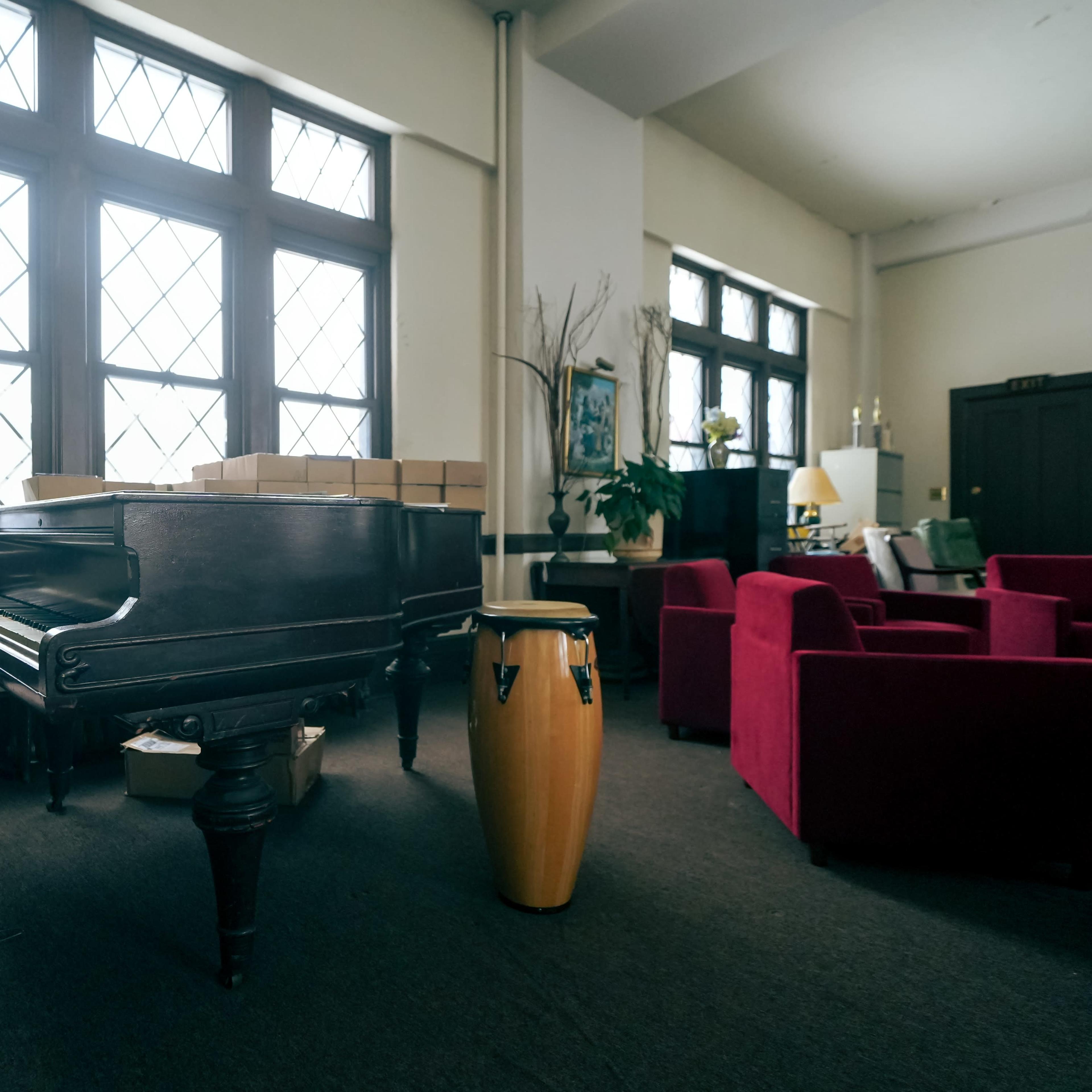 A wooden grand piano and a conga drum are positioned beside a set of red armchairs in a spacious room with large windows.