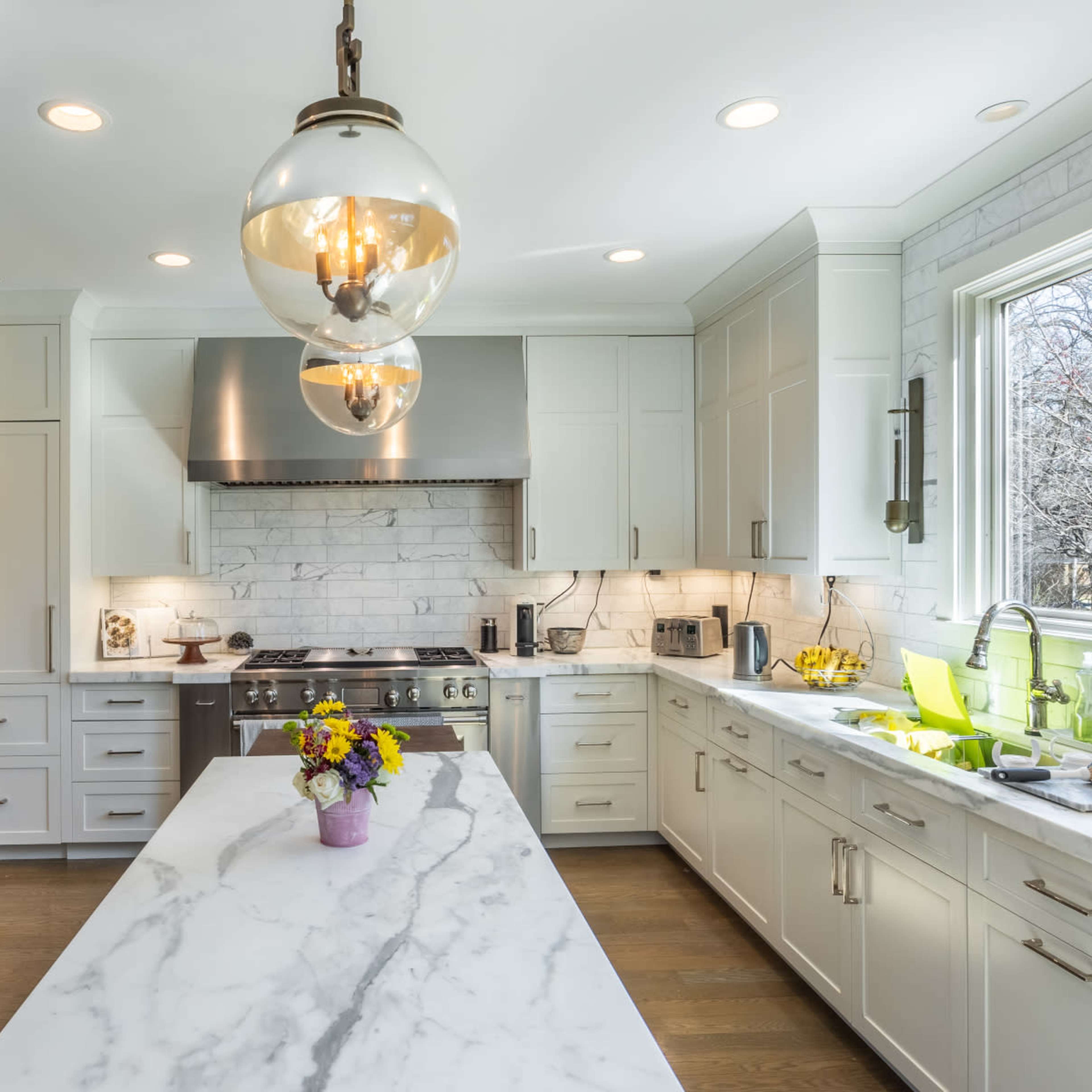 A modern kitchen features a large marble island, dark wood cabinets, stainless steel appliances, and a window overlooking a garden.