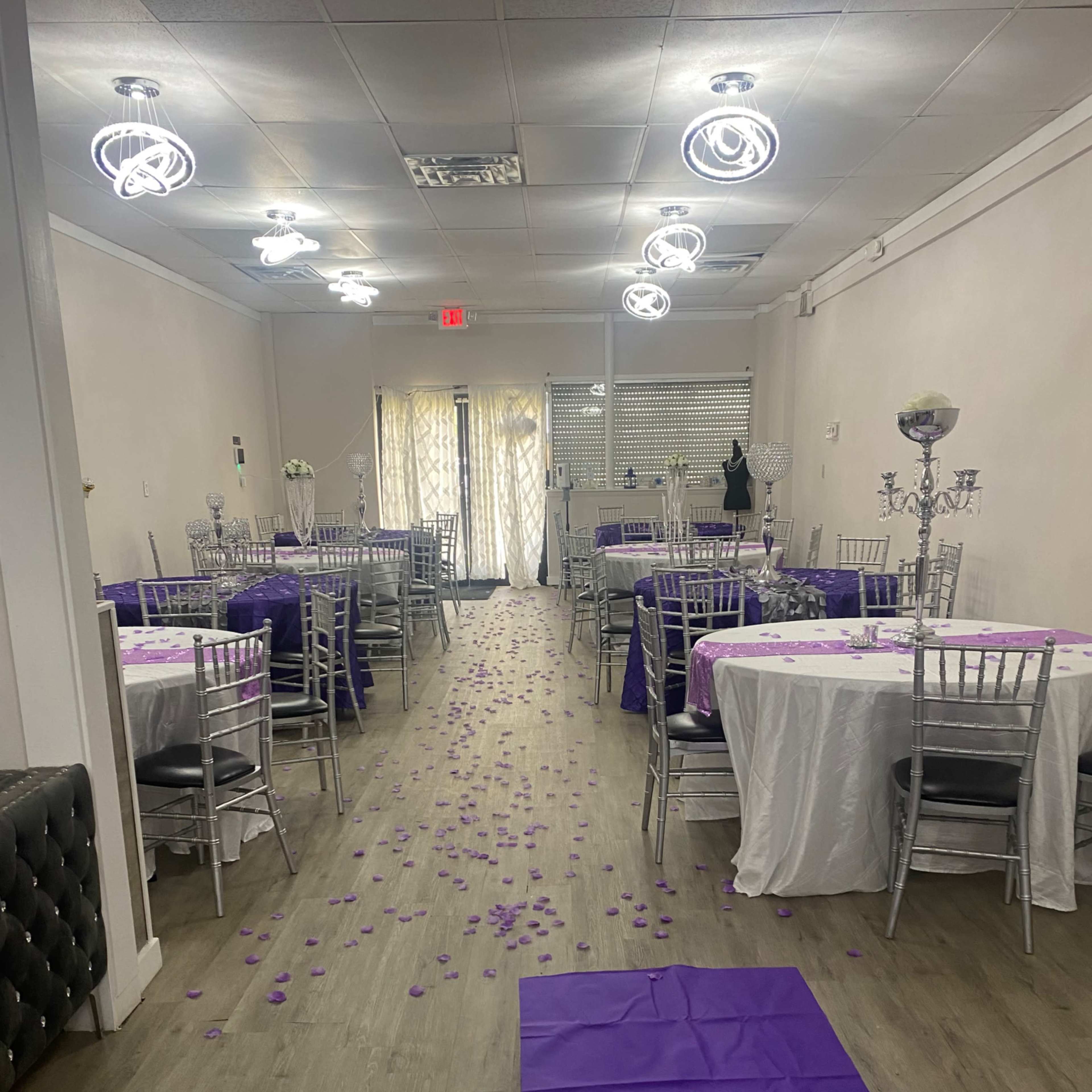 A banquet hall set up with circular tables covered in white and purple tablecloths, featuring silver chairs and a path of purple petals leading down the center.