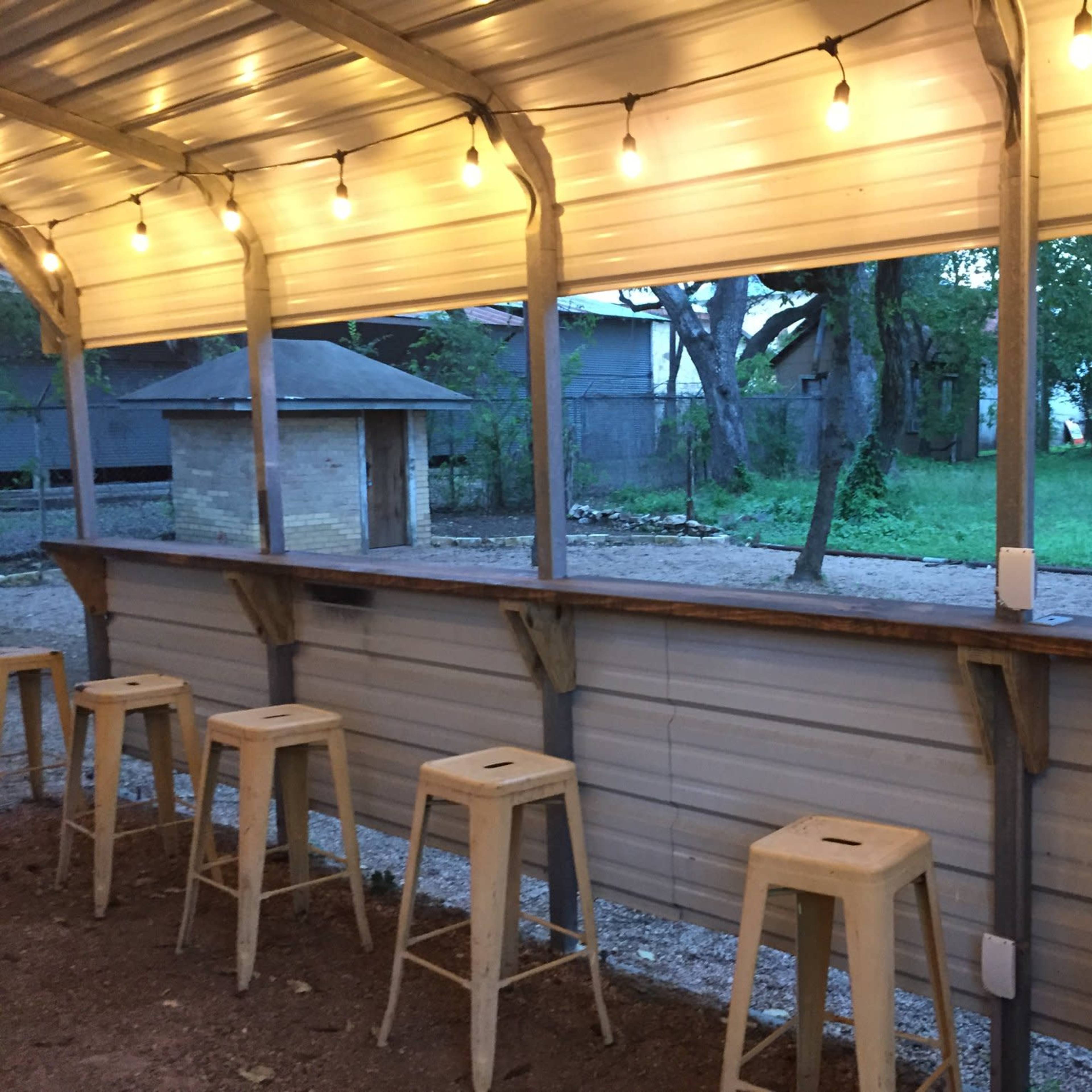 A covered outdoor bar area with wooden stools, string lights overhead, and an open view of a grassy yard bordered by trees and buildings.