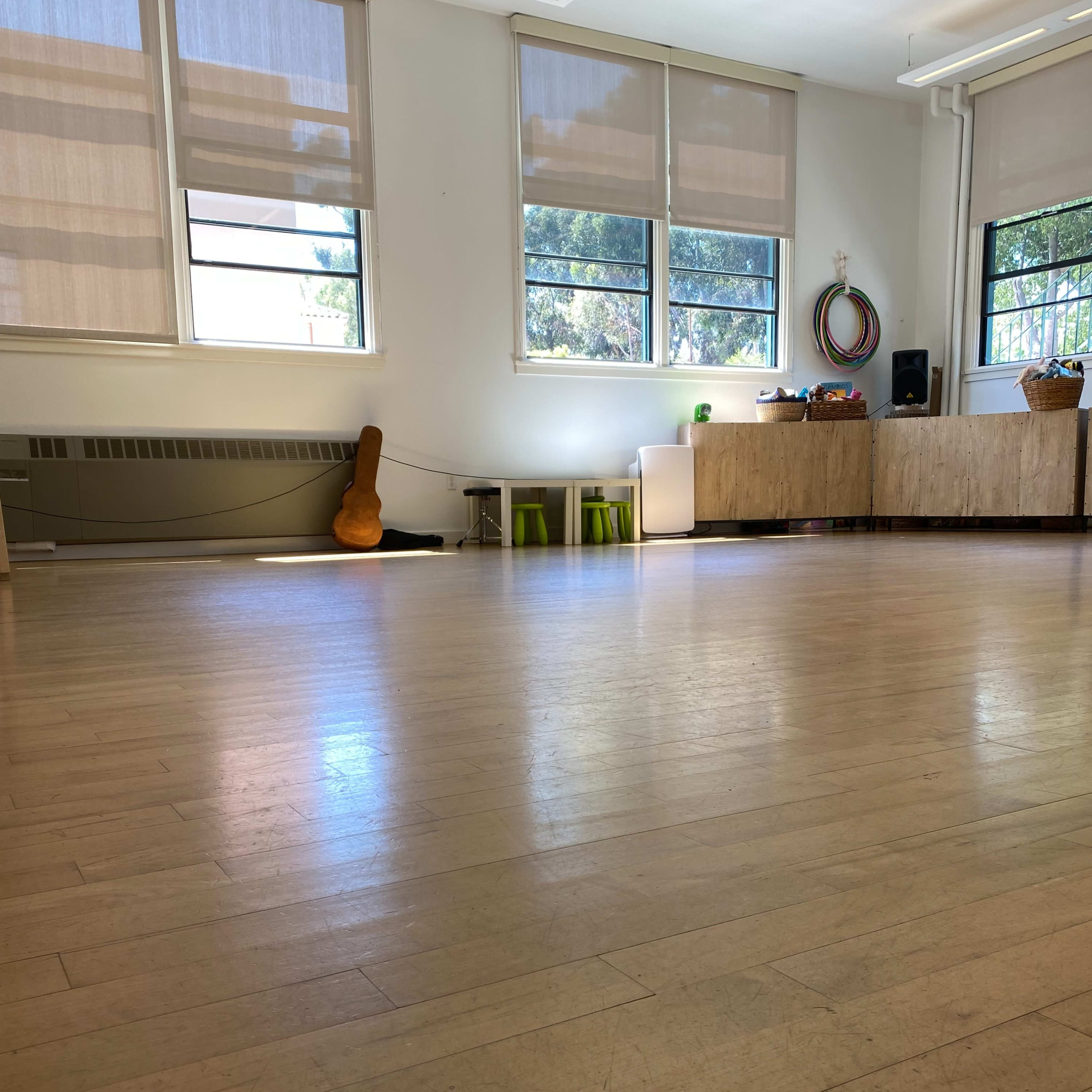 A spacious room with wooden floors, large windows, and minimal furniture including a guitar and some green stools.