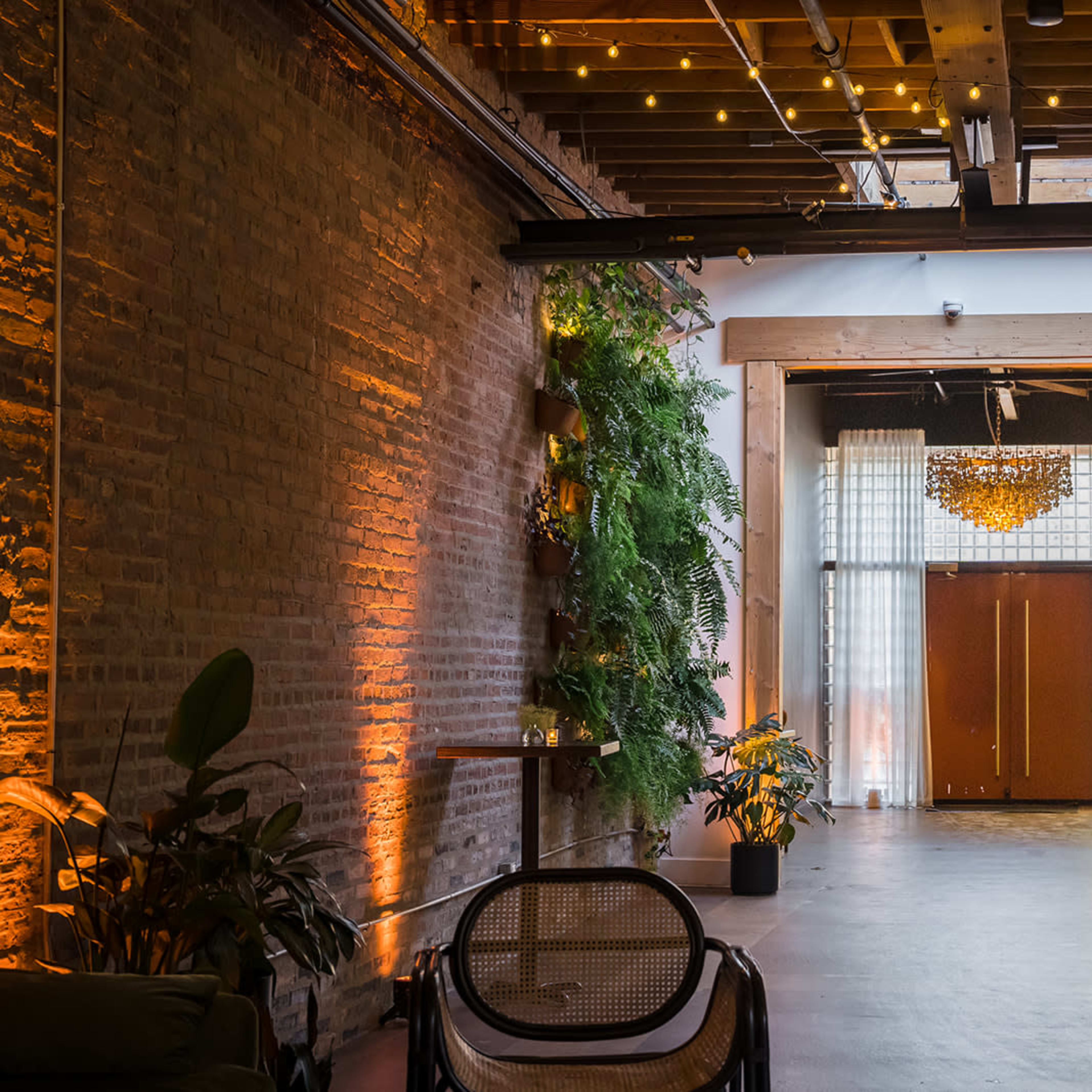 A spacious interior with exposed brick walls, wooden beams, and plants lining the entrance, leading to a set of large wooden doors illuminated by a chandelier overhead.