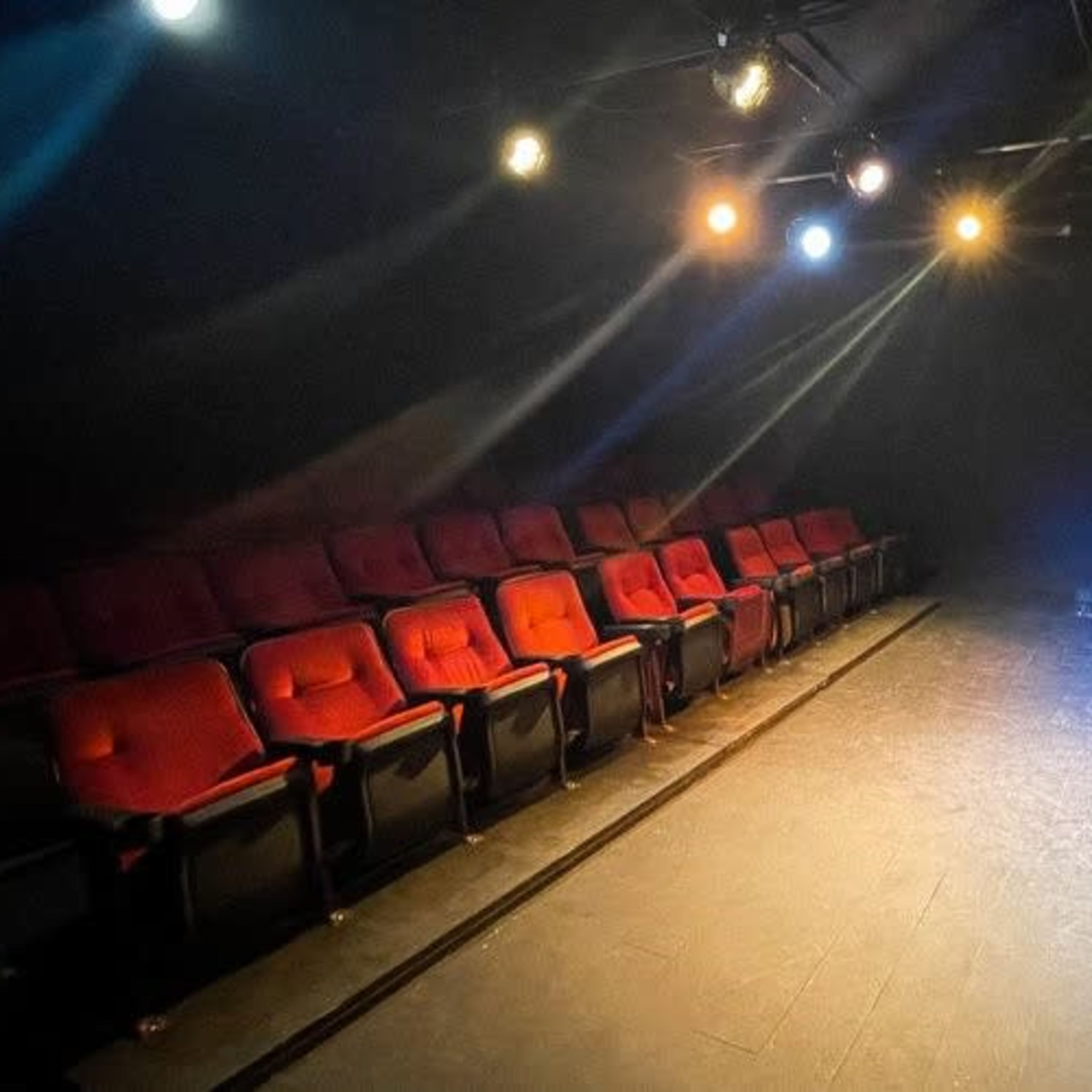 A dimly lit theater features rows of red seats facing a dark stage, illuminated by overhead lights.