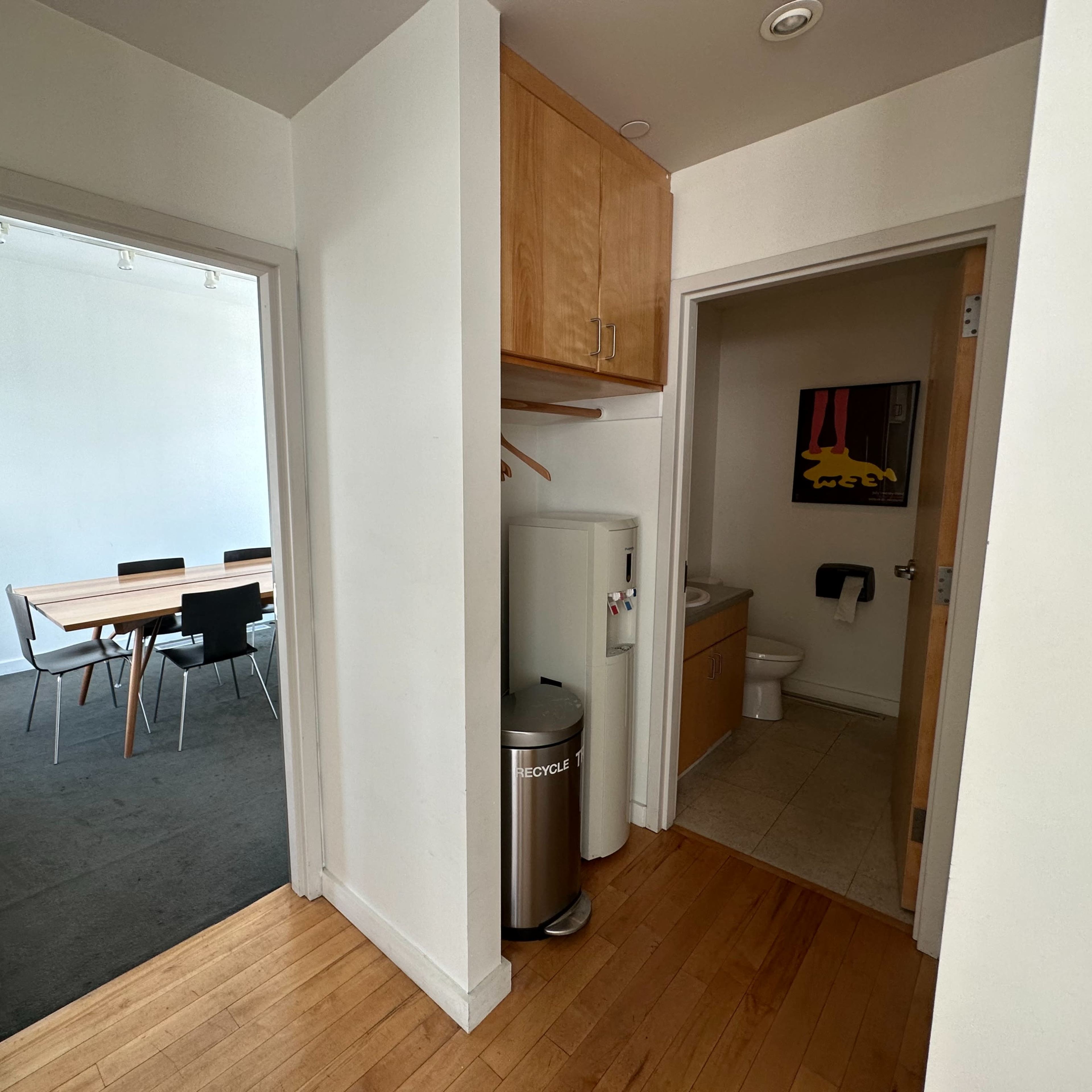 A hallway with two doorways leading to a meeting room on the left and a bathroom on the right, featuring a refrigerator and a trash can near the bathroom entrance.