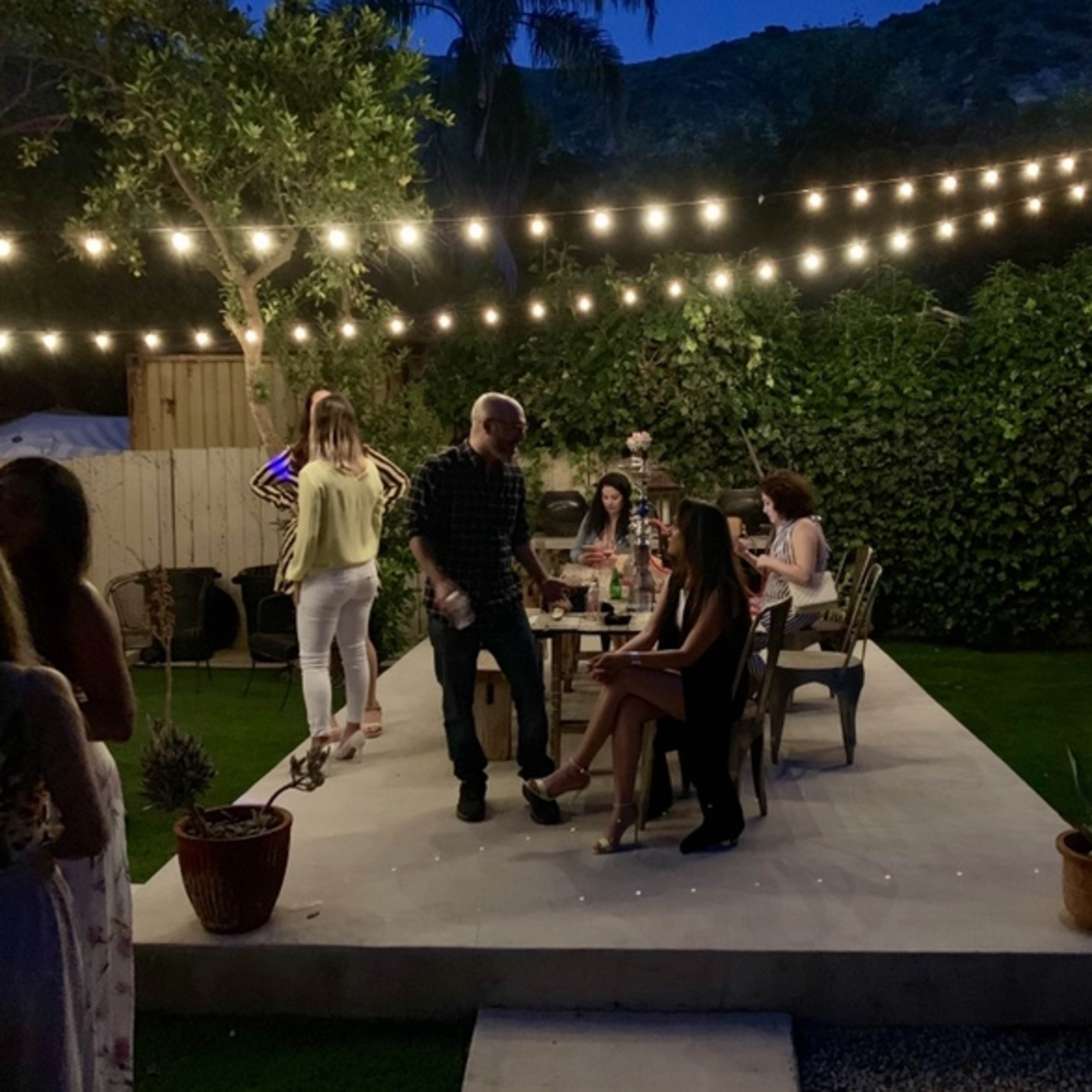 A group of people socialize in a backyard setting under string lights, surrounded by greenery and potted plants.