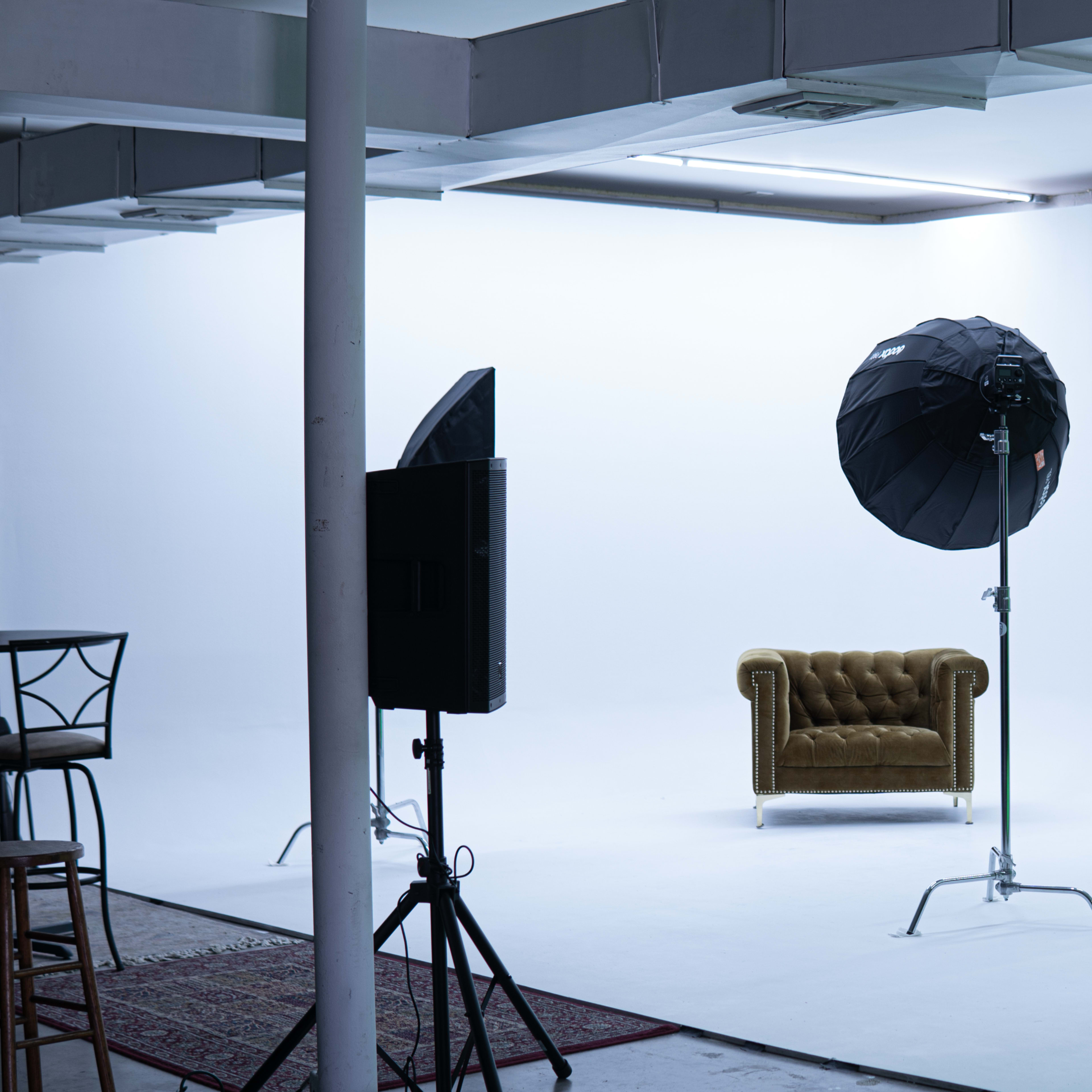 A photography studio features a white backdrop, a brown tufted armchair, and two large softbox lights on stands.