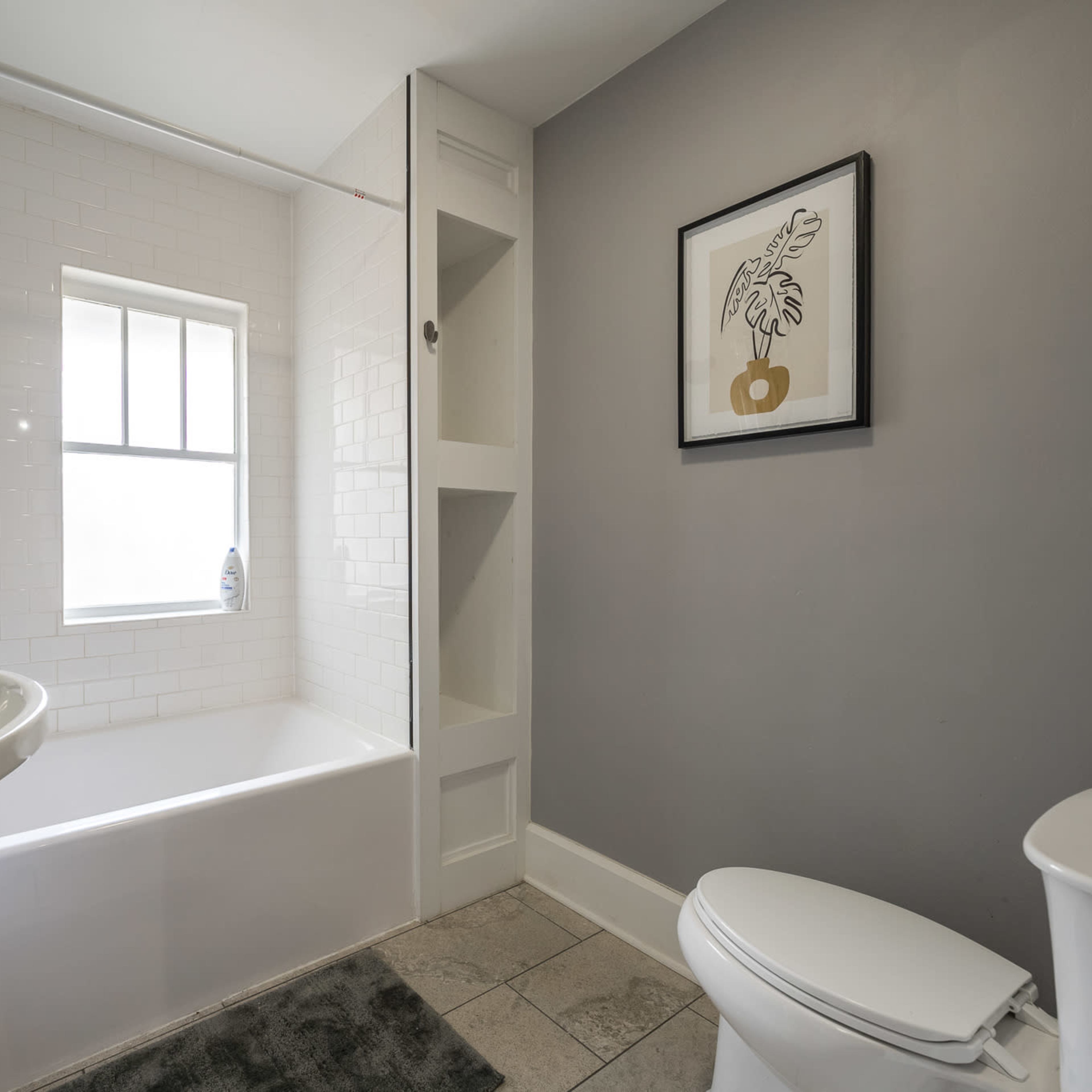 A modern bathroom featuring a bathtub with a shower, a pedestal sink, a toilet, and a window, all set against gray walls and a patterned shower curtain.