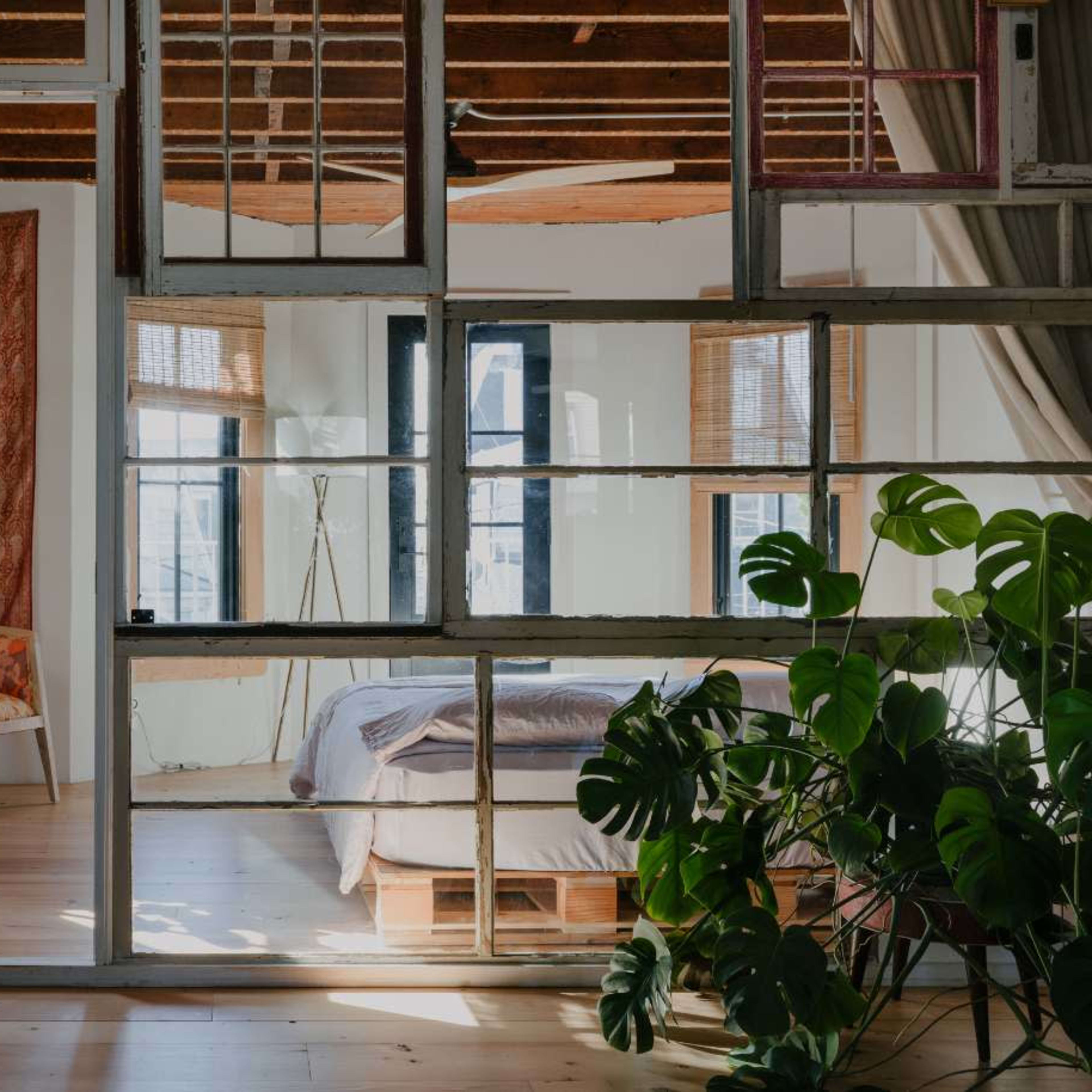 A bright, airy bedroom with large windows, a plant in the foreground, and a cozy seating area visible through a glass partition.