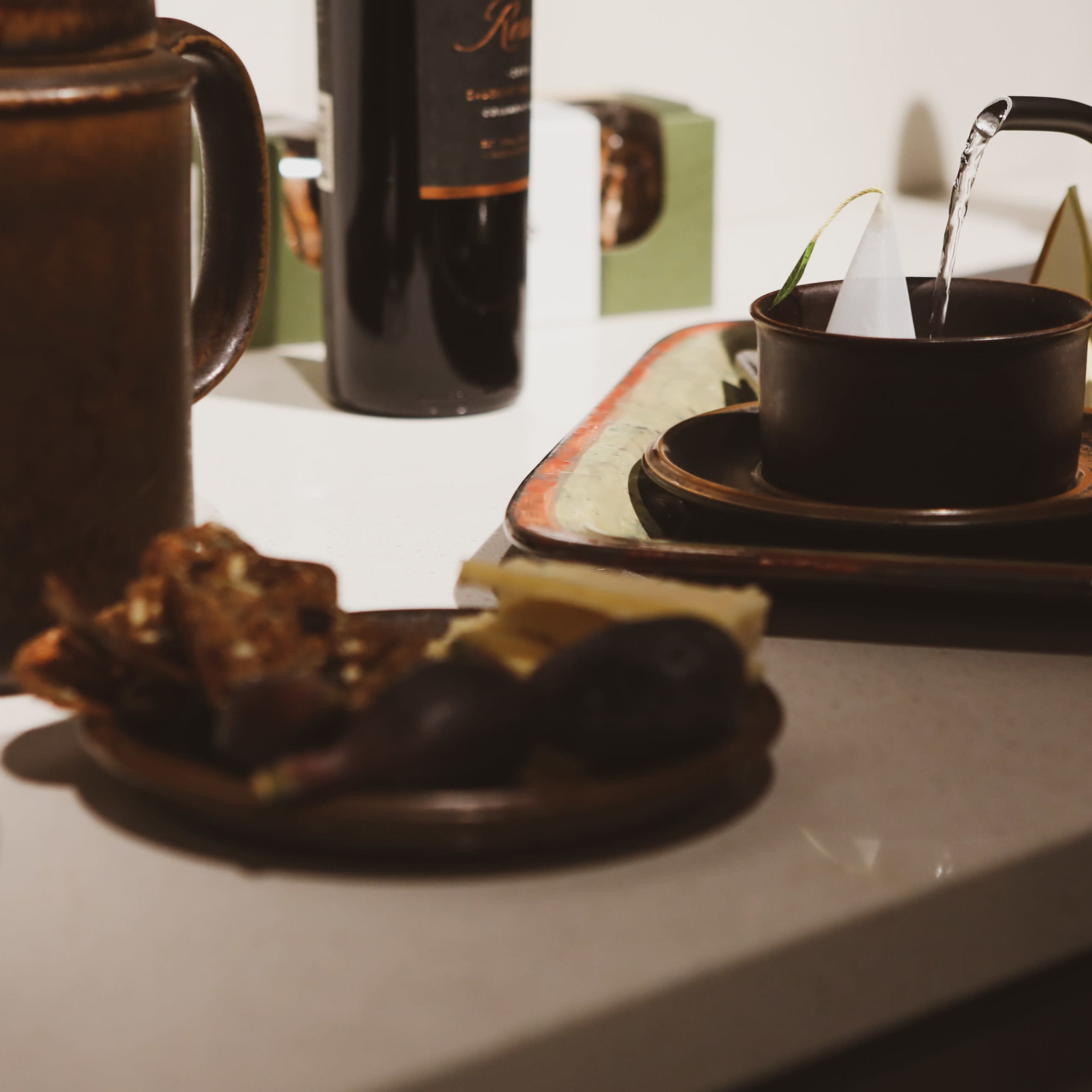 A teapot pours water into a dark ceramic cup placed on a decorative tray, alongside a plate of assorted fruits and snacks.