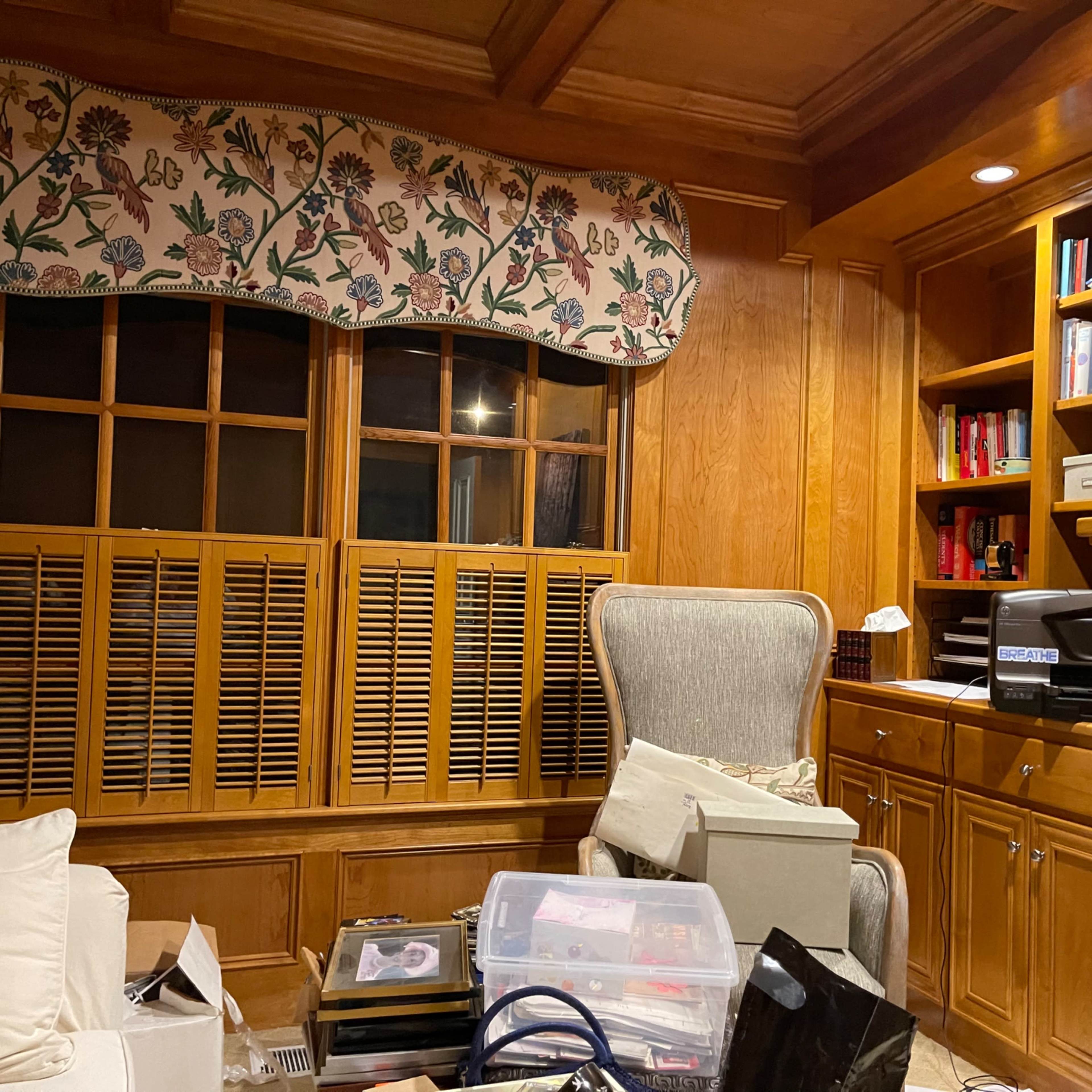A cluttered room with wooden paneling, a large armchair, a window with a patterned valance, and shelves filled with books and boxes.