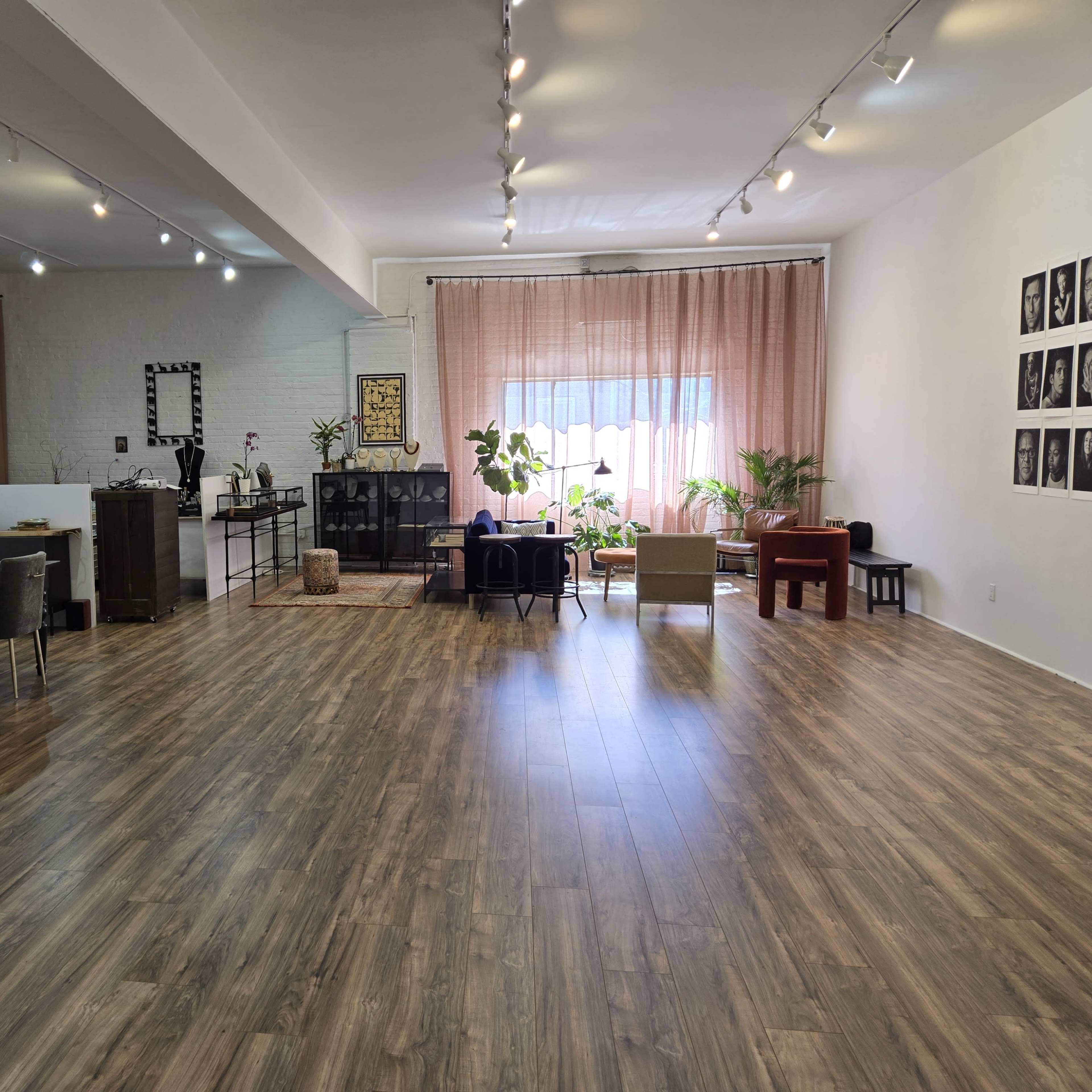 A spacious room with wooden flooring, equipped with a dining table, chairs, plants, and a large window draped with curtains.