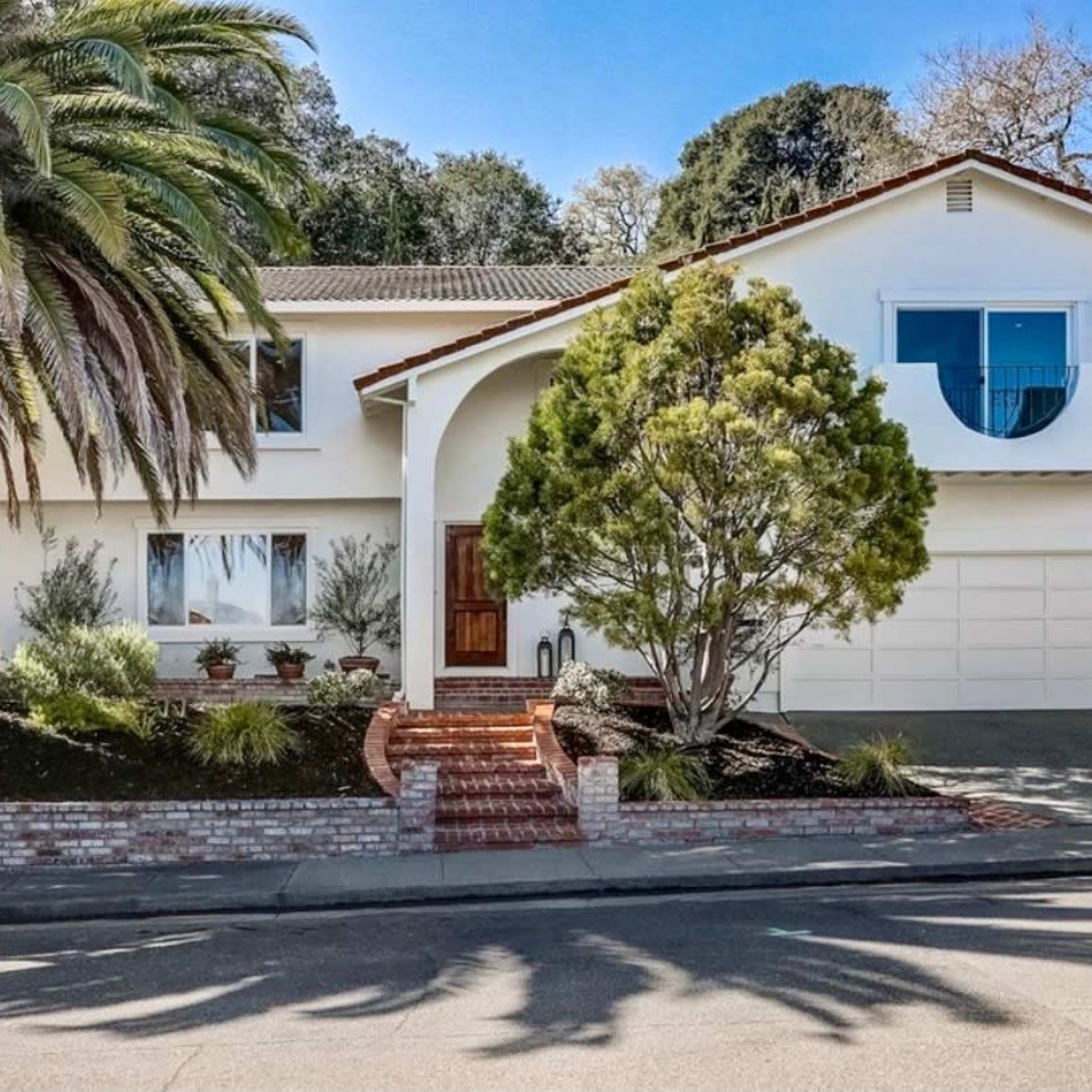 A two-story house with a palm tree and a circular window, situated on a sloped lot with a brick walkway leading to the entrance.