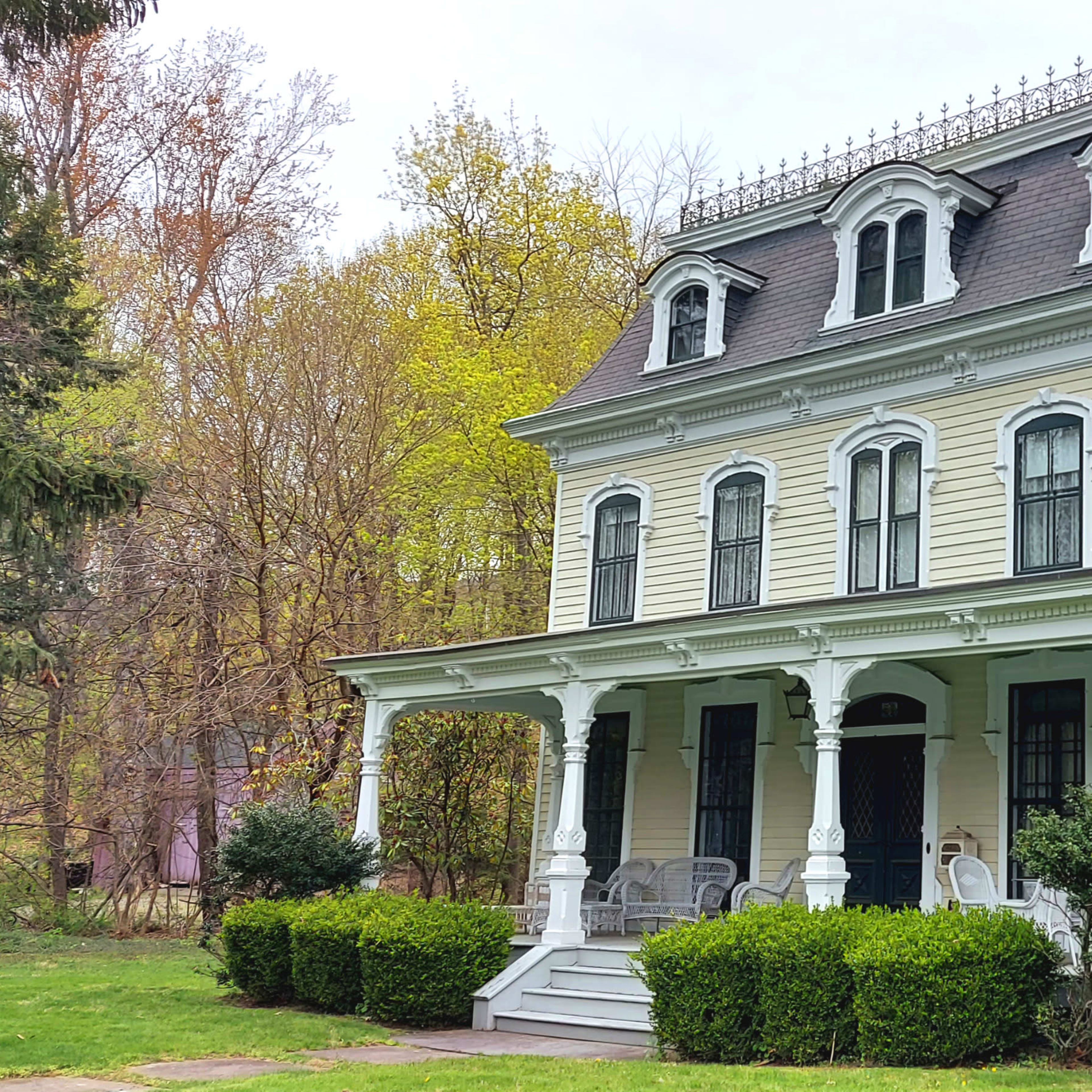 A large, three-story Victorian-style house with a wraparound porch and intricate roof detailing, surrounded by greenery.