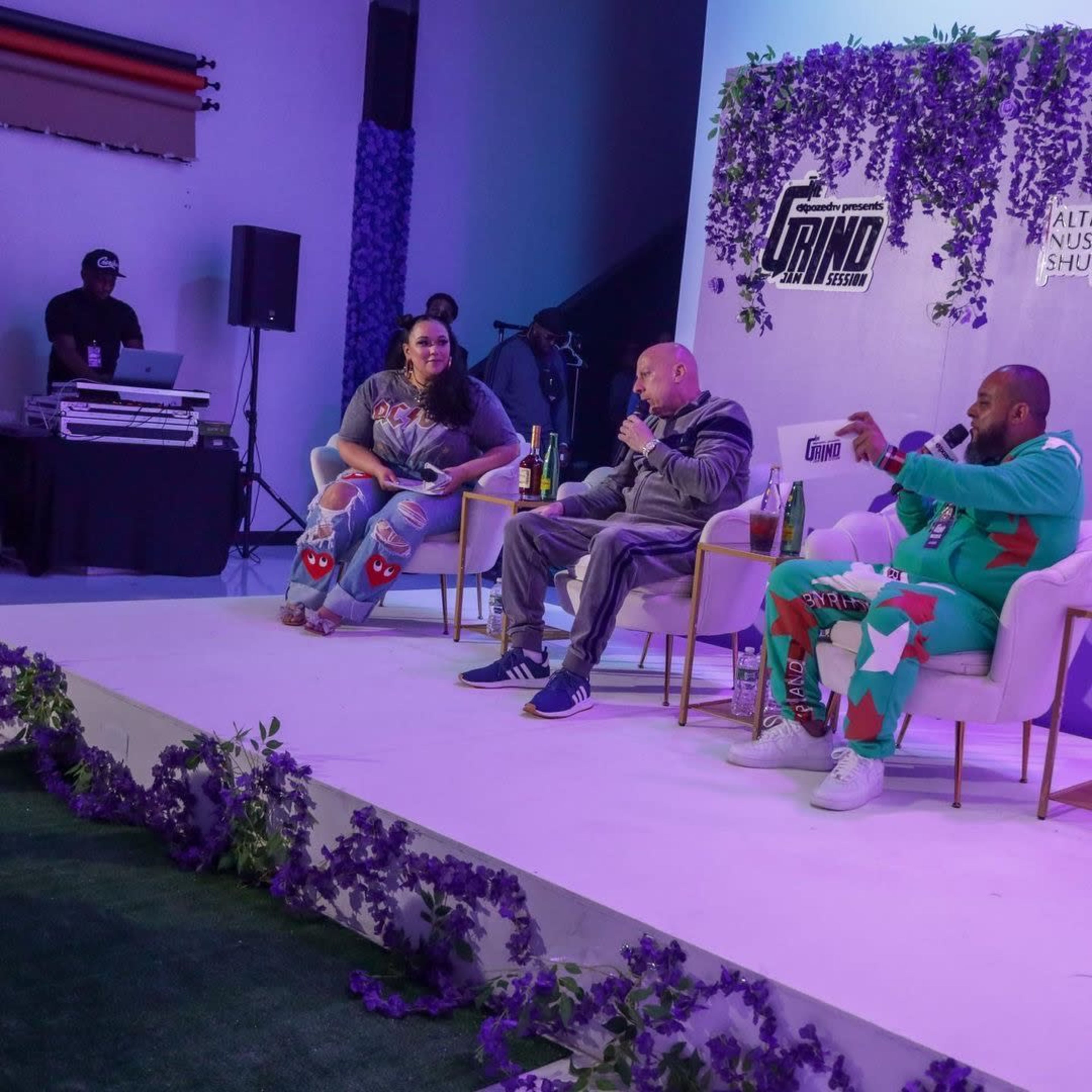 A panel discussion takes place on stage, with three individuals seated and a DJ setup visible in the background.