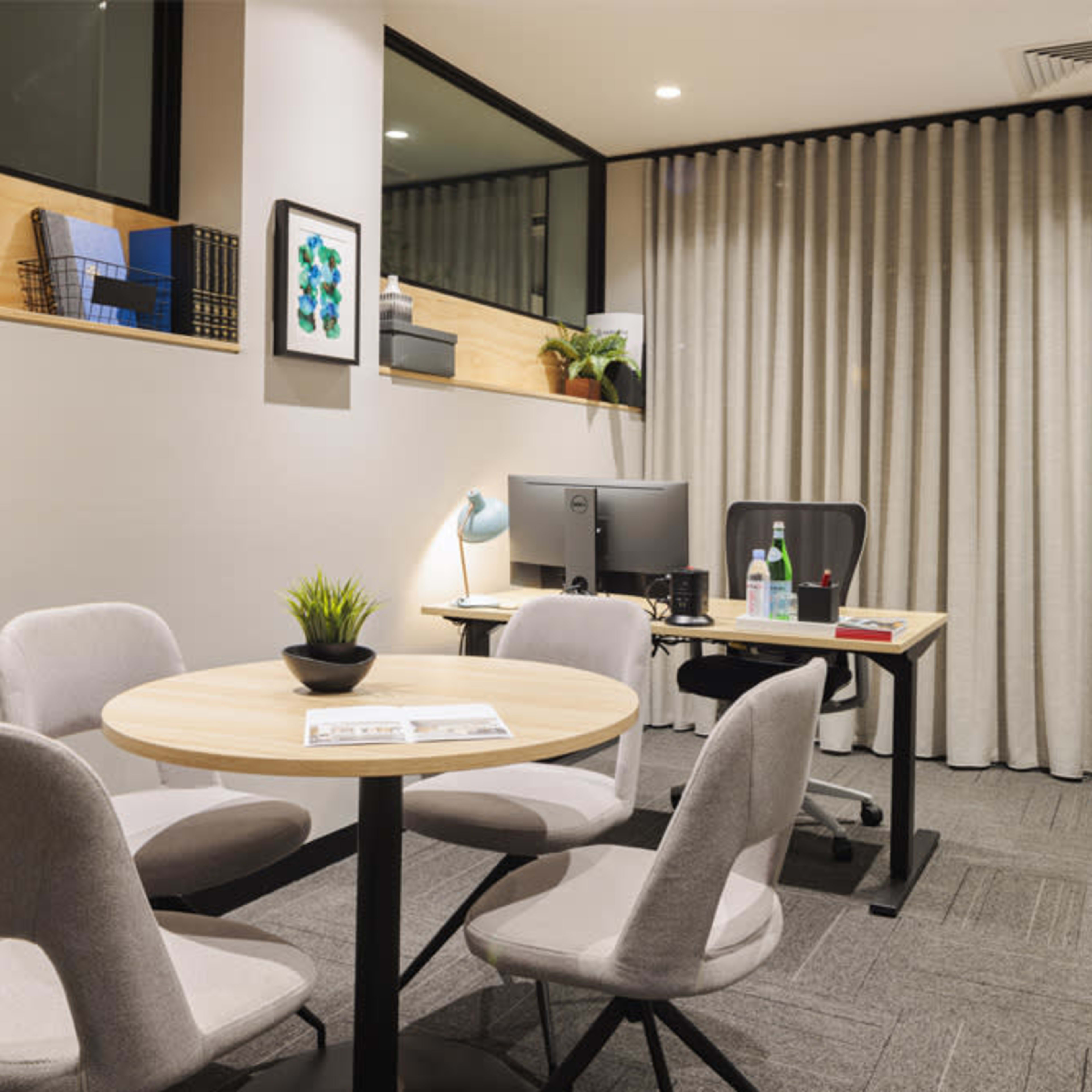 A modern office space featuring a round table with four chairs, a computer desk with a monitor, and a presentation board against a backdrop of curtains.