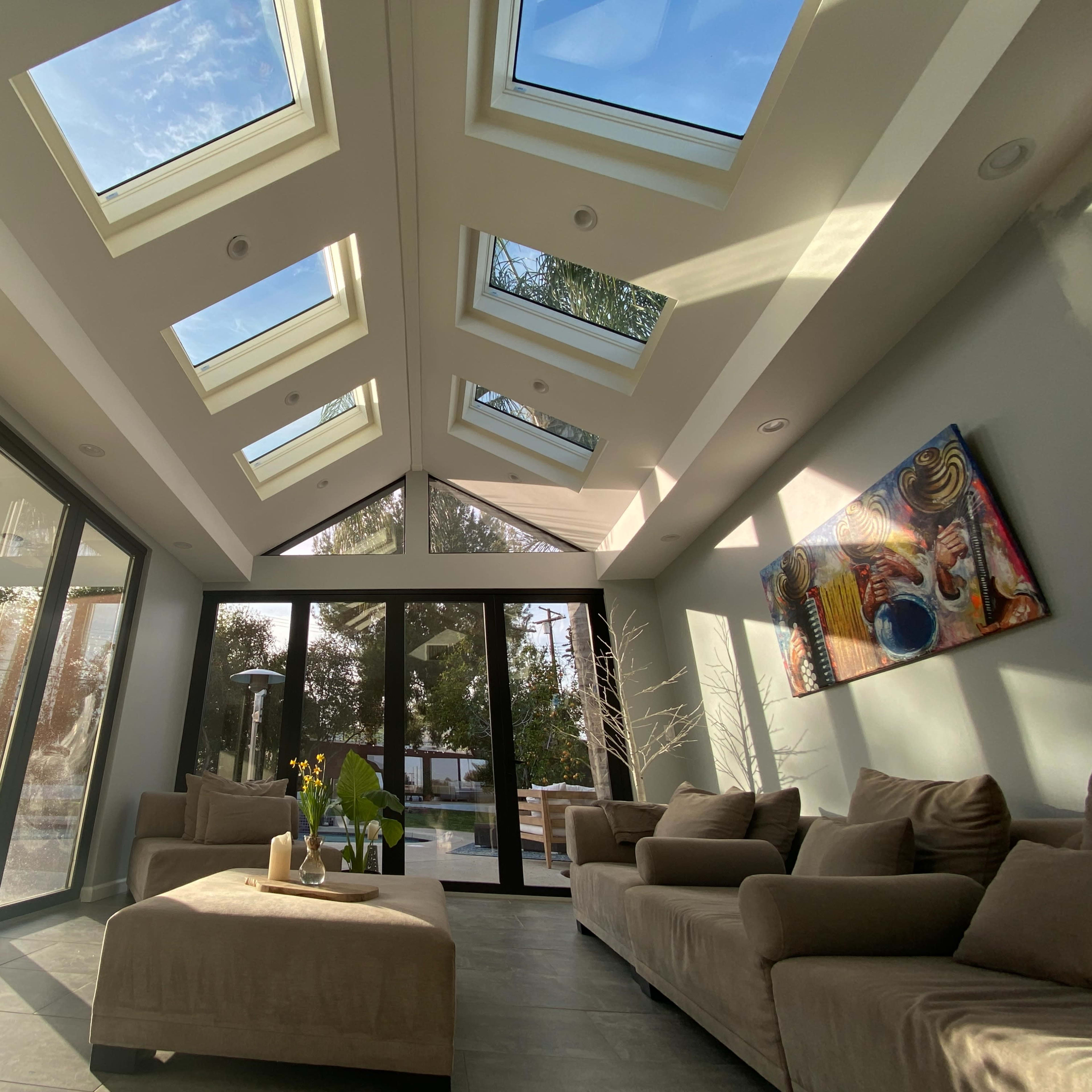 A sunlit modern living room with large skylights and glass walls, featuring beige sofas and a decorative painting on the wall.