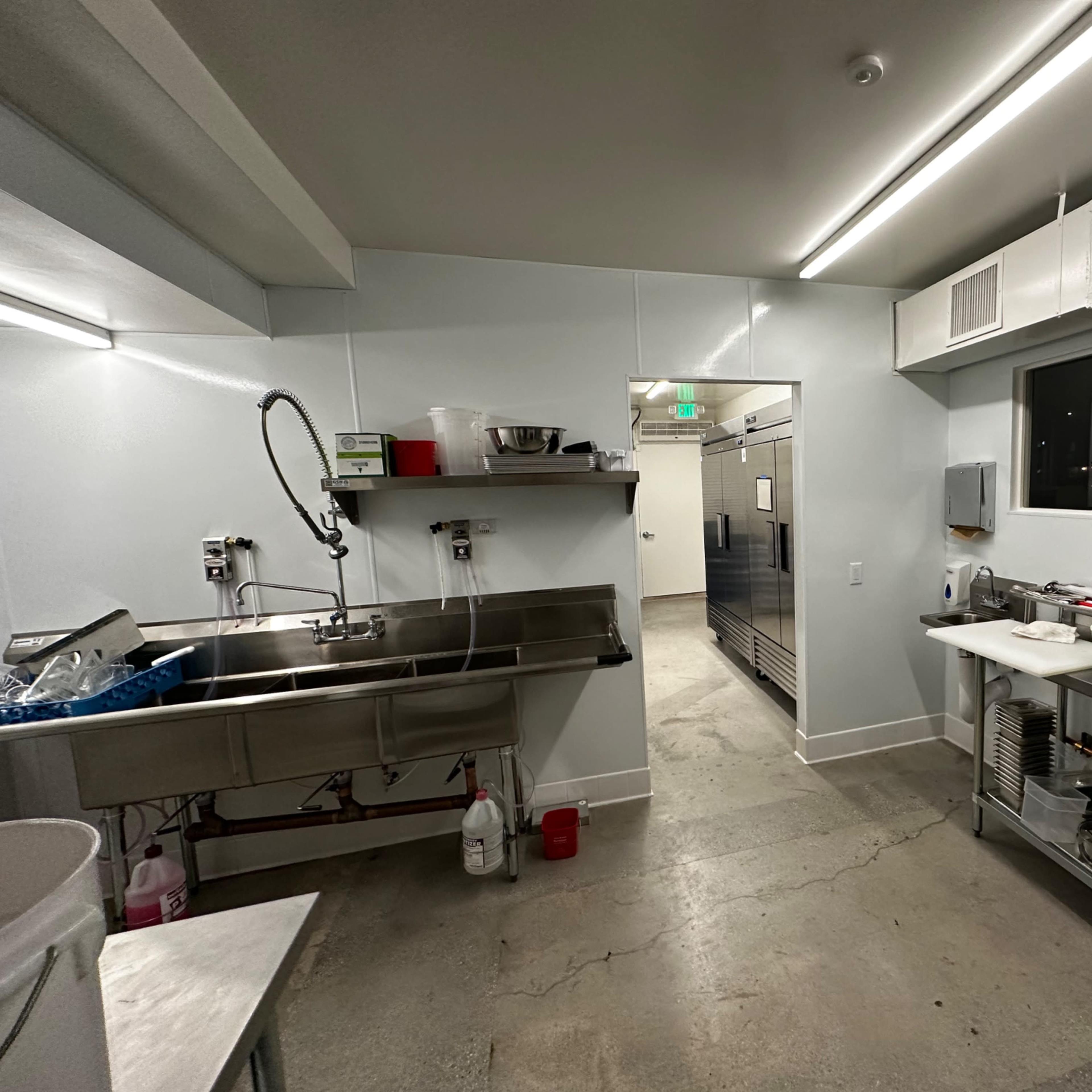 A clean commercial kitchen featuring stainless steel countertops, a sink, and a large refrigerator in the background.