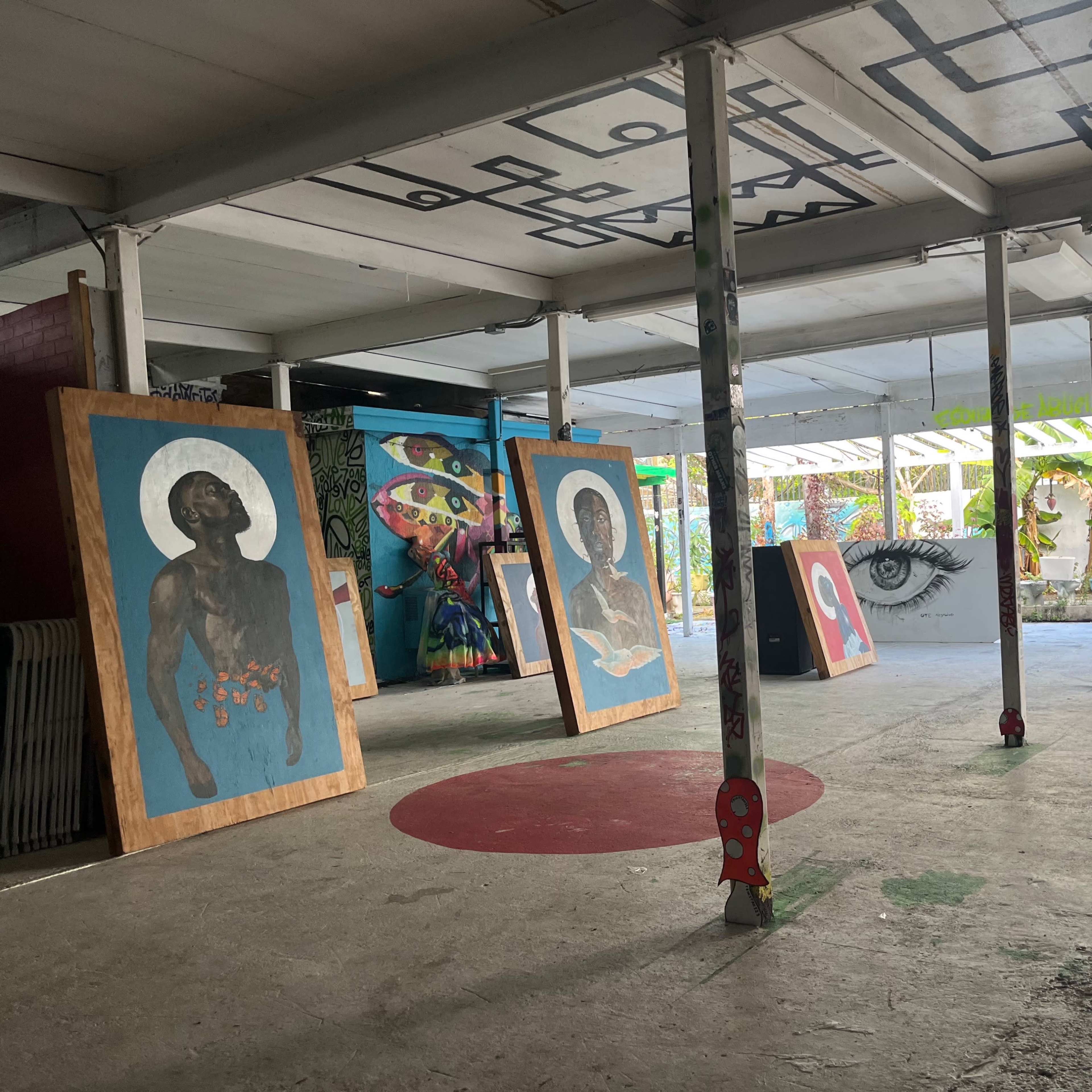 An indoor space featuring large, colorful artwork displayed against a backdrop of vibrant murals and greenery visible through open doors.
