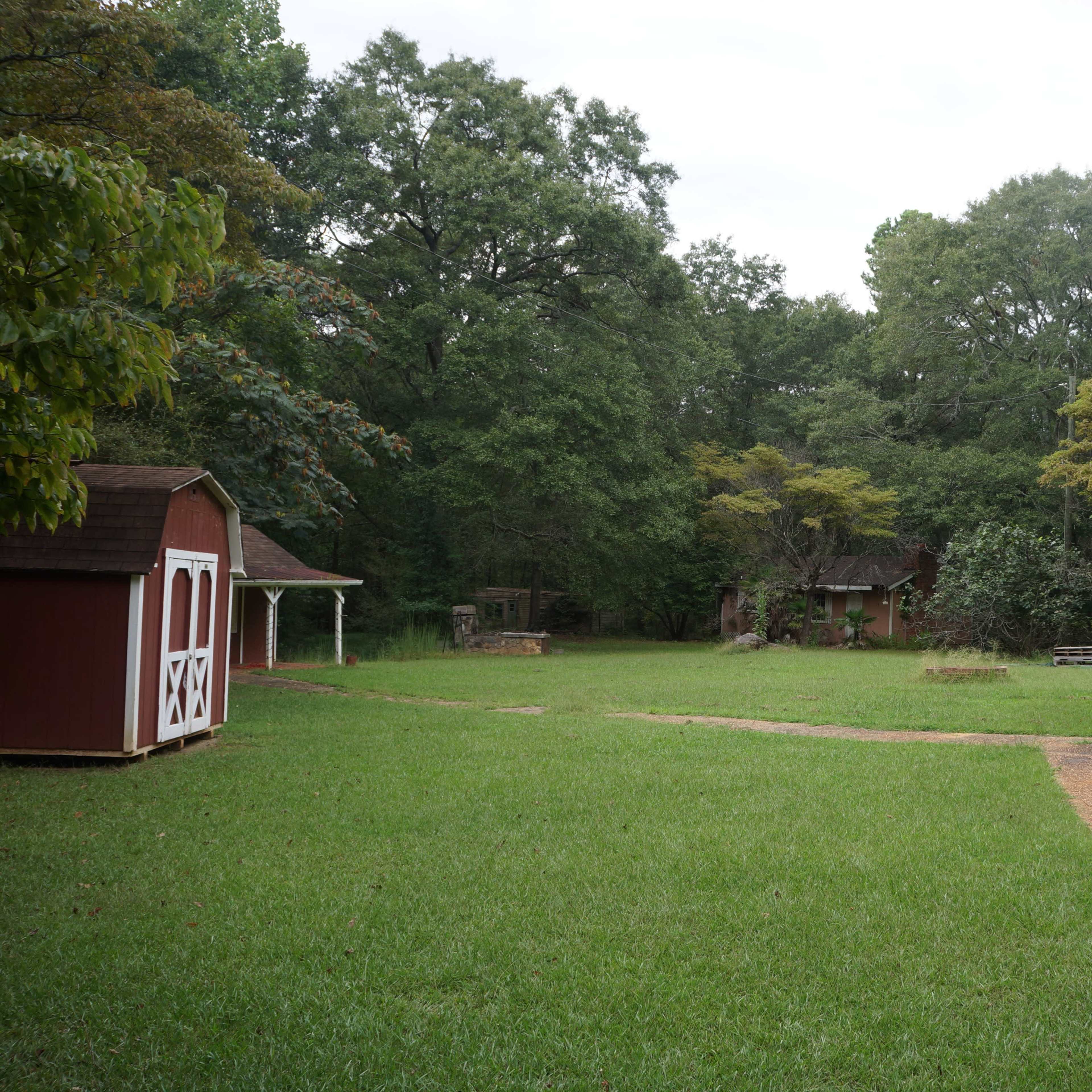 A green yard with a gravel path leading towards wooden structures and surrounded by trees.
