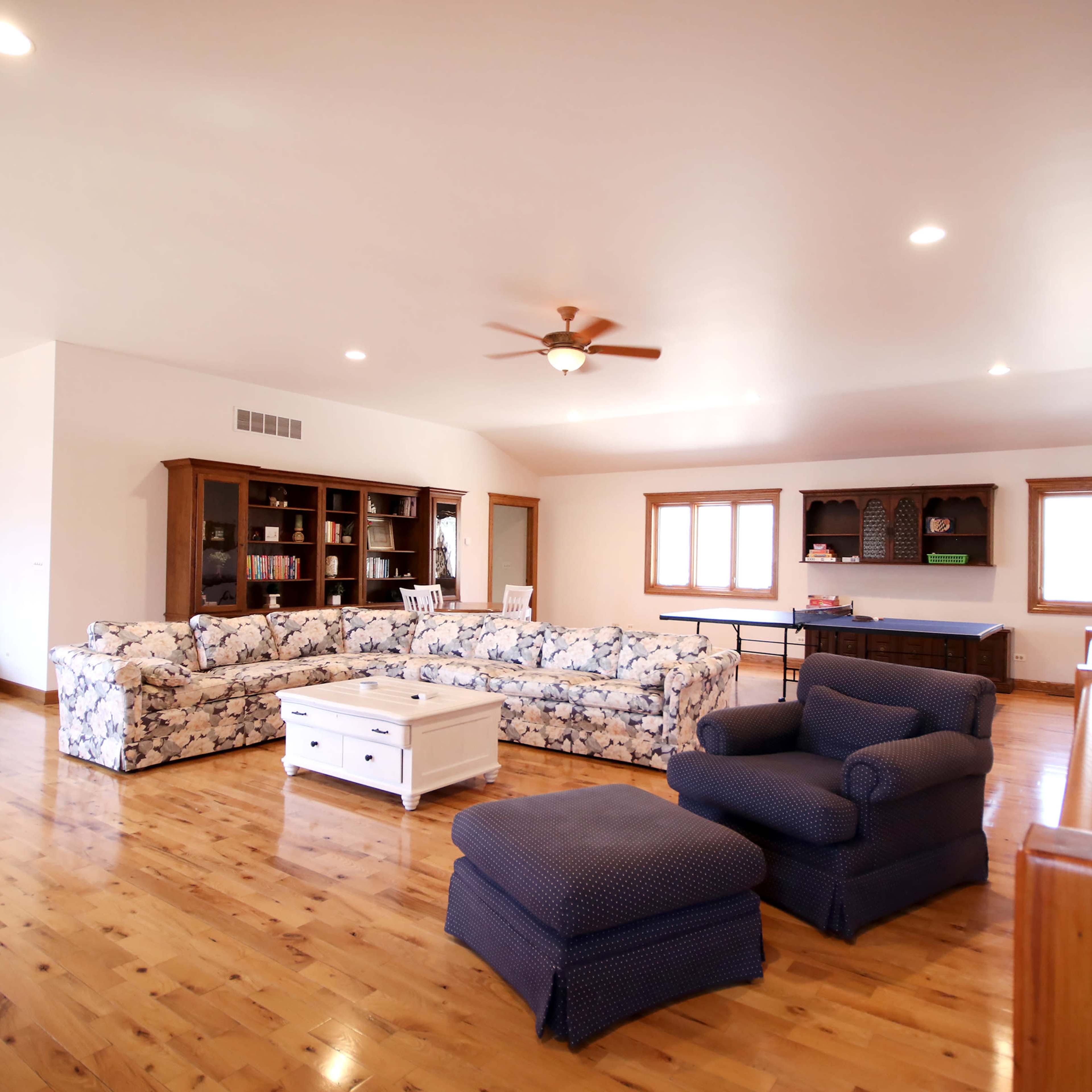 The spacious living room features a floral-patterned sectional sofa, a wooden bookcase, and a ceiling fan, all set against polished hardwood floors.