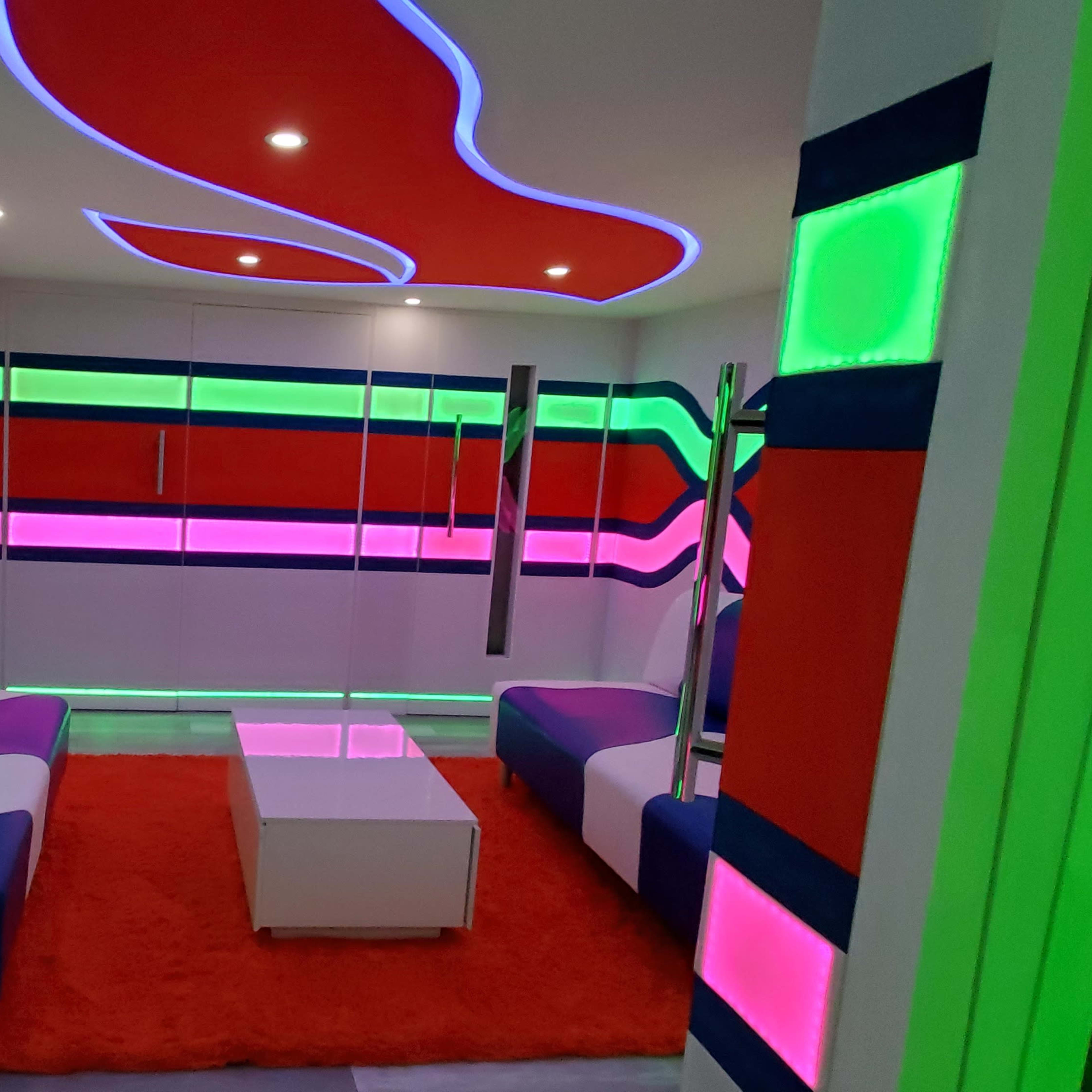 A modern living room with vibrant, multicolored wall designs and furniture featuring white and colored couches, a glass coffee table, and illuminated accents.