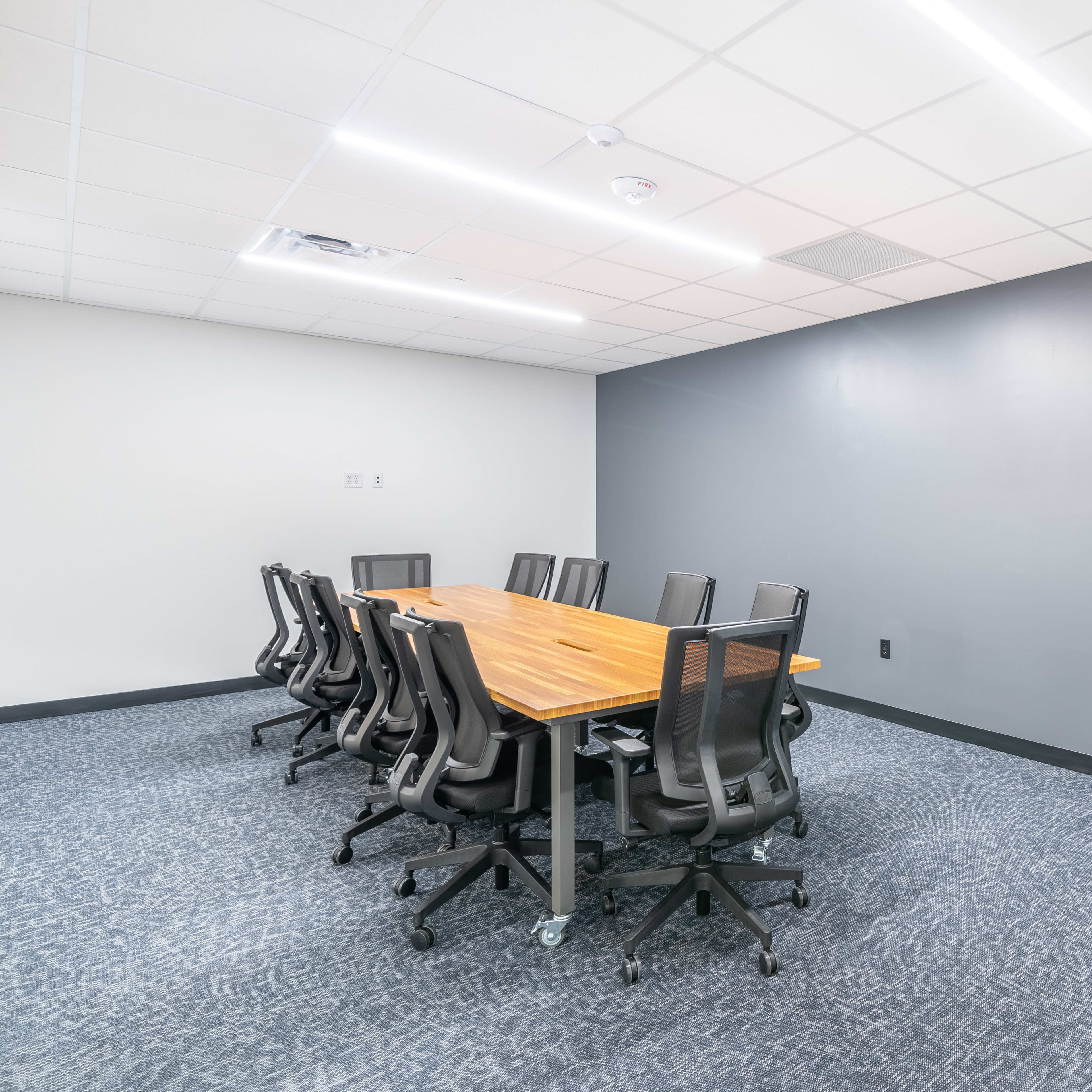 A conference room features a large wooden table surrounded by ergonomic black office chairs and plain gray walls.