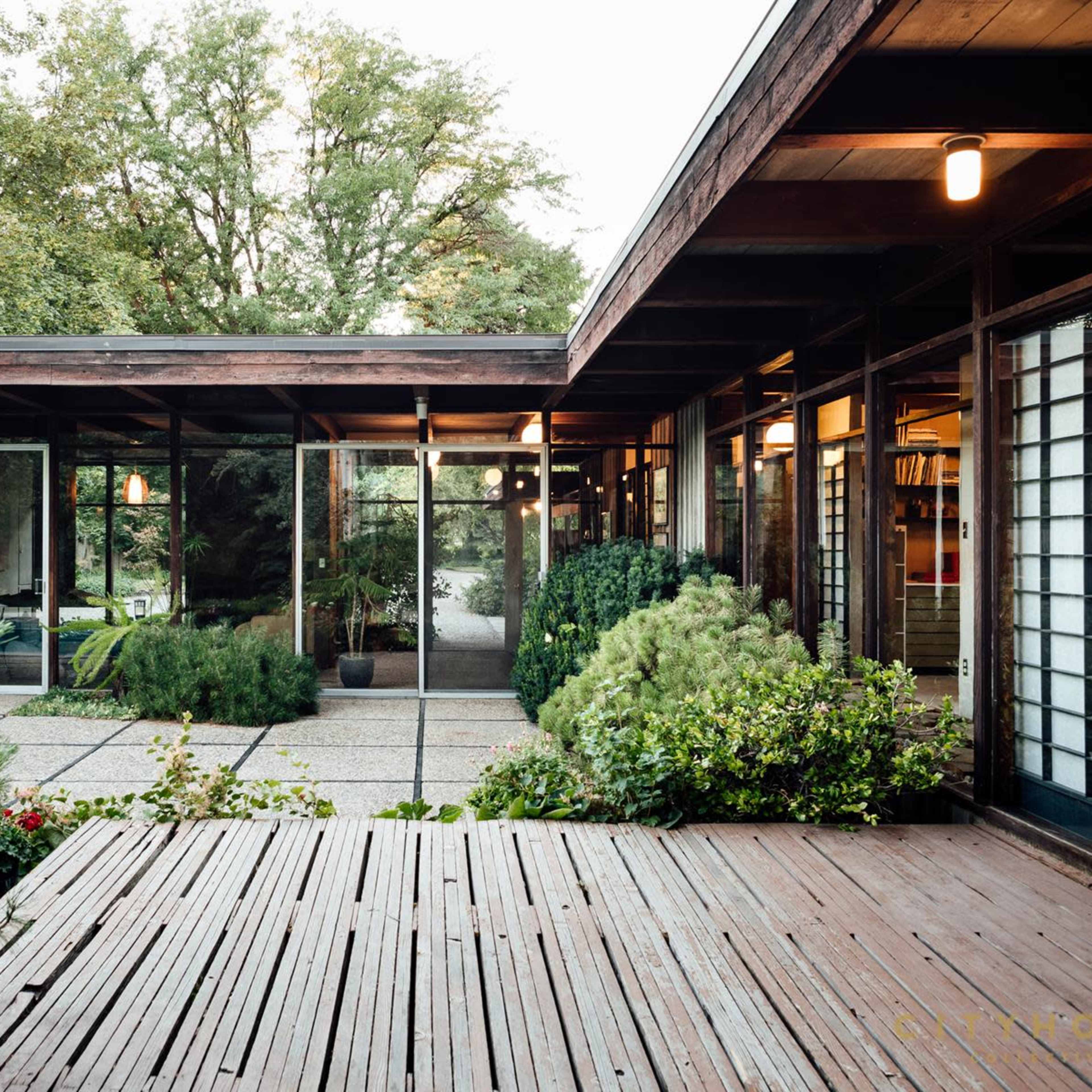 A modern, glass-enclosed house surrounded by greenery and a wooden deck.