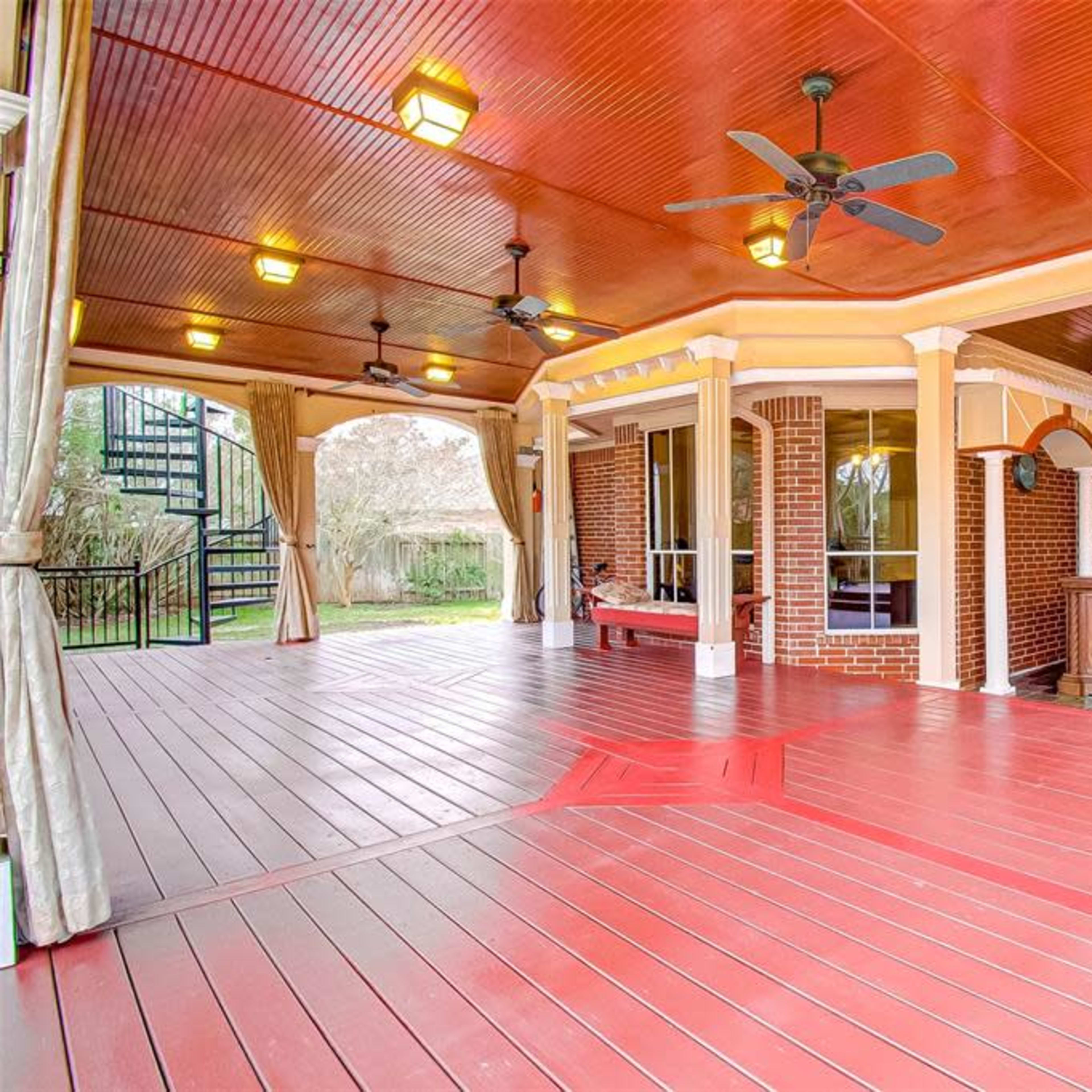 A spacious covered patio with wooden flooring, ceiling fans, and brick columns, featuring a spiral staircase leading to an upper level.
