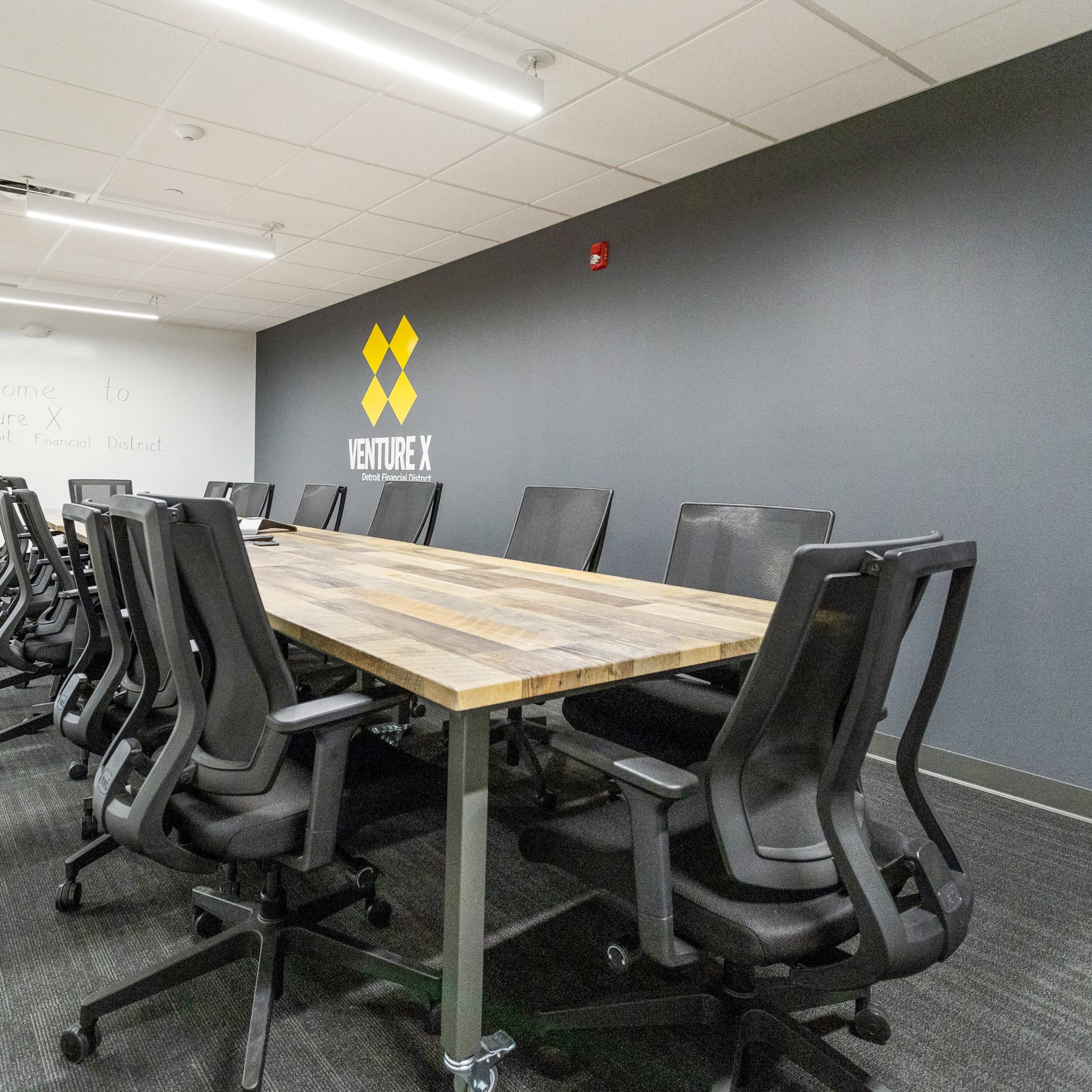 A modern conference room features a long wooden table surrounded by black ergonomic chairs, with a large logo on the gray wall.