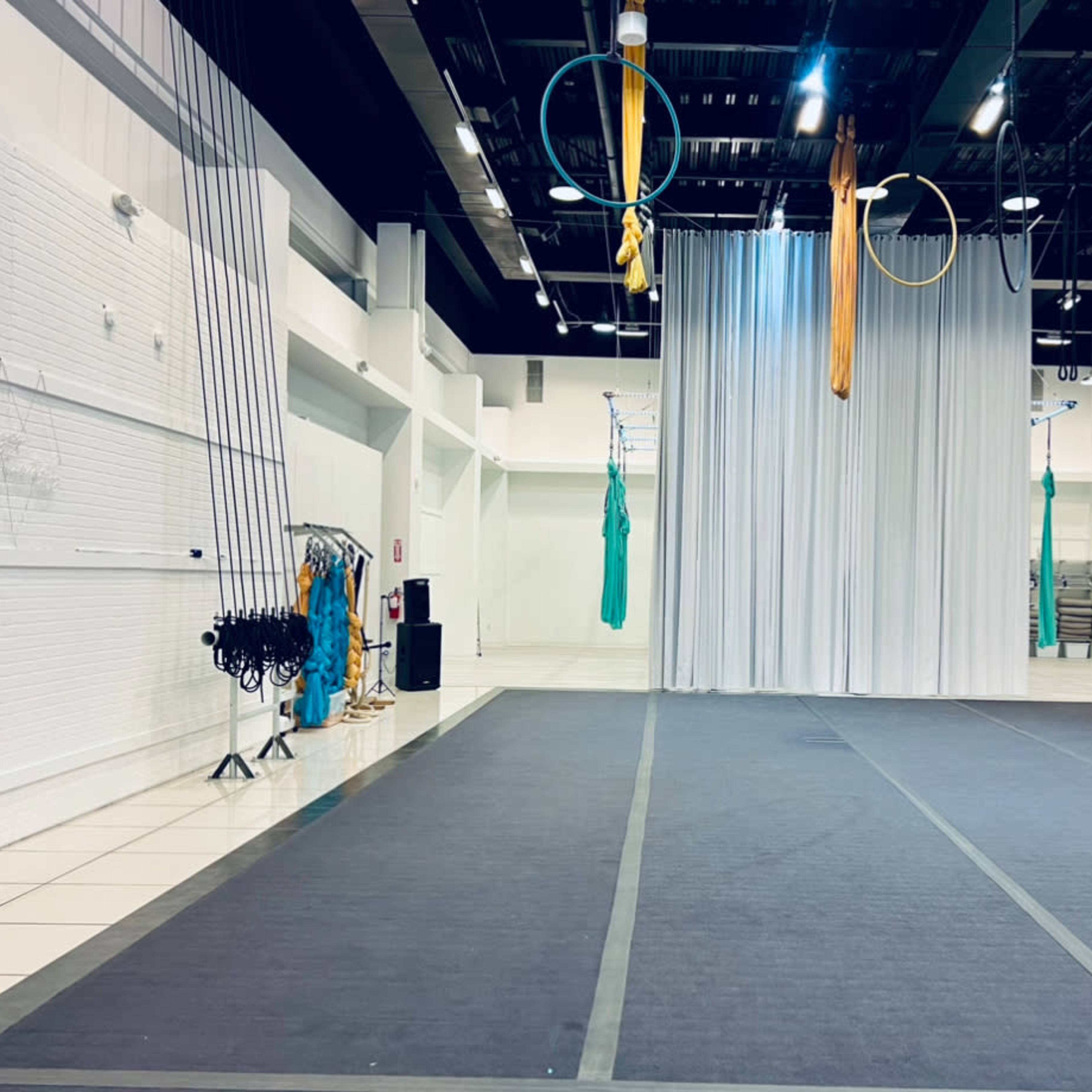 A spacious indoor area set up for aerial arts, featuring silks, hoops, and a tall fabric curtain, along with equipment like a scaffolding and musical speakers.