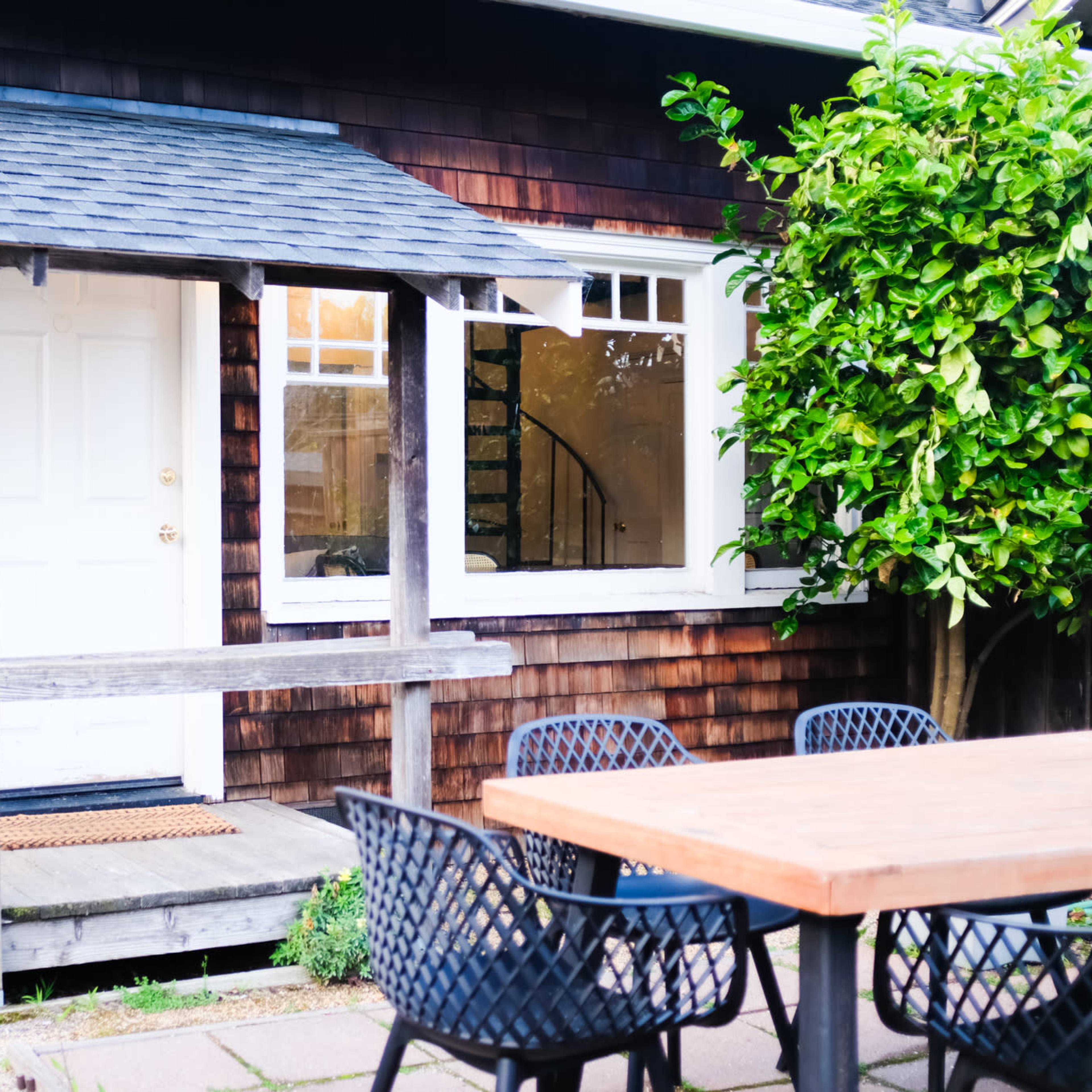 A small wooden porch with a white door, adjacent to a building with dark wooden siding, and a table with black chairs positioned on a patio.
