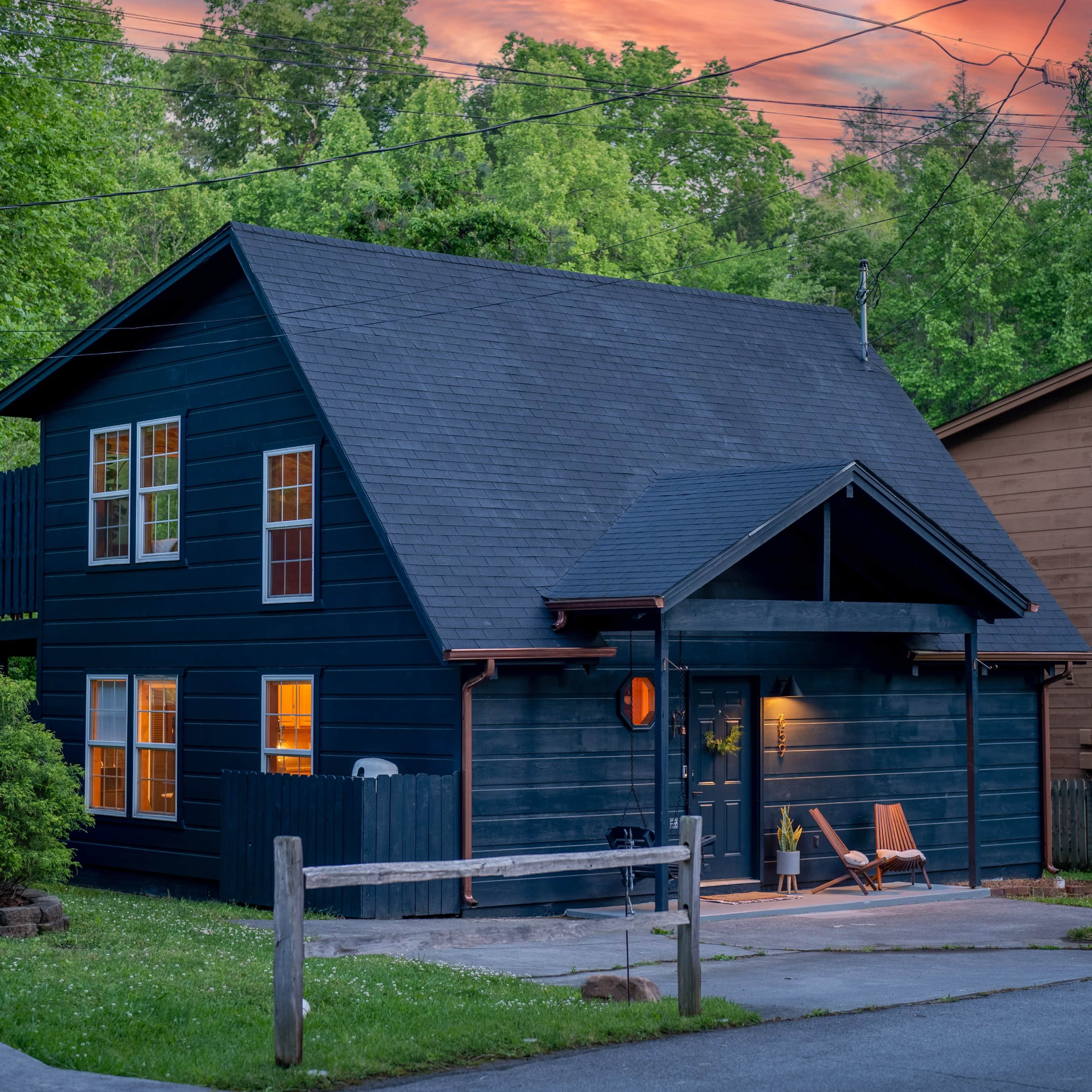 A dark-painted house with a sloped roof is situated on a serene street, illuminated by the warm glow of sunset.