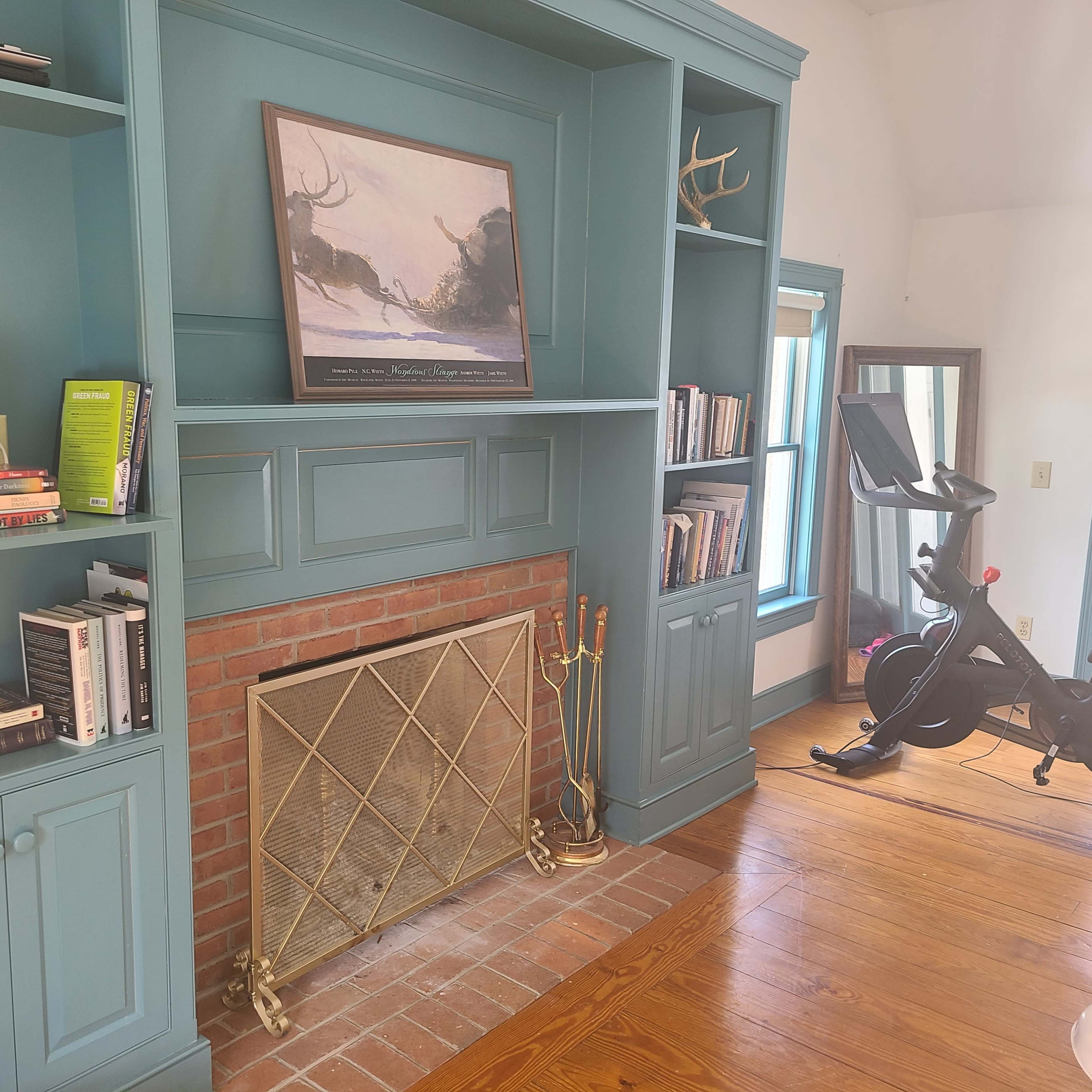 A room with a built-in bookshelf, a brick fireplace, a mounted painting, and an exercise bike.
