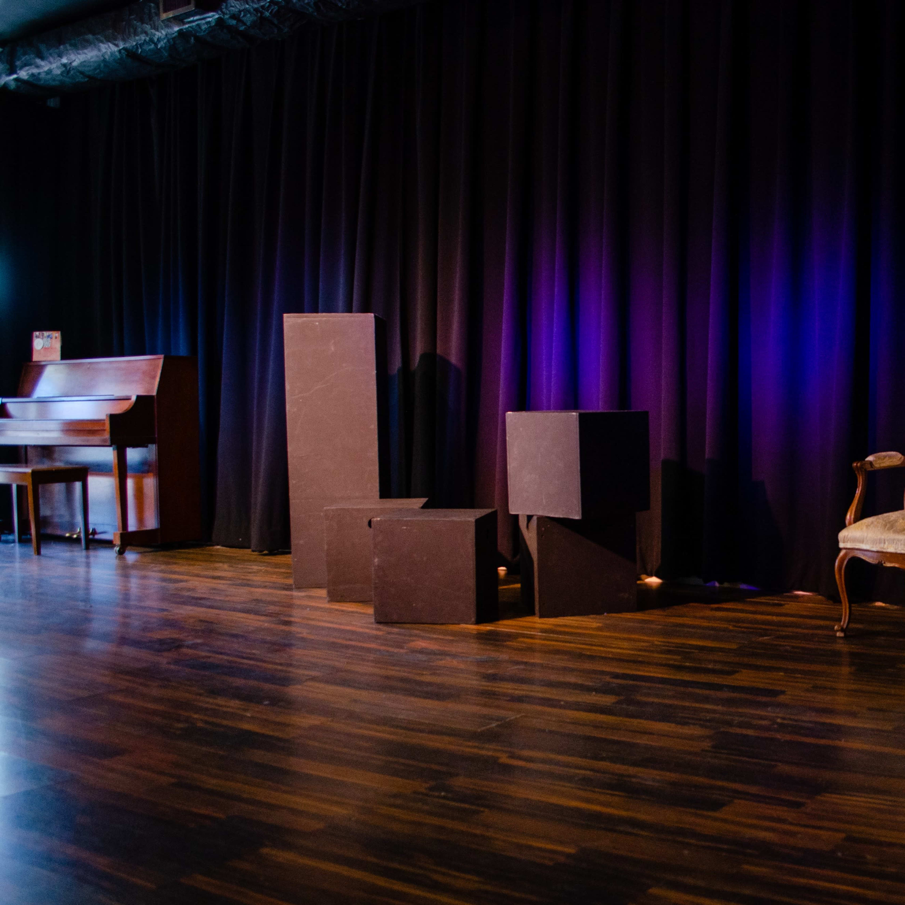 A stage is set with a black curtain in the background, a vintage chair on one side, and various black geometric shapes scattered across the wooden floor.