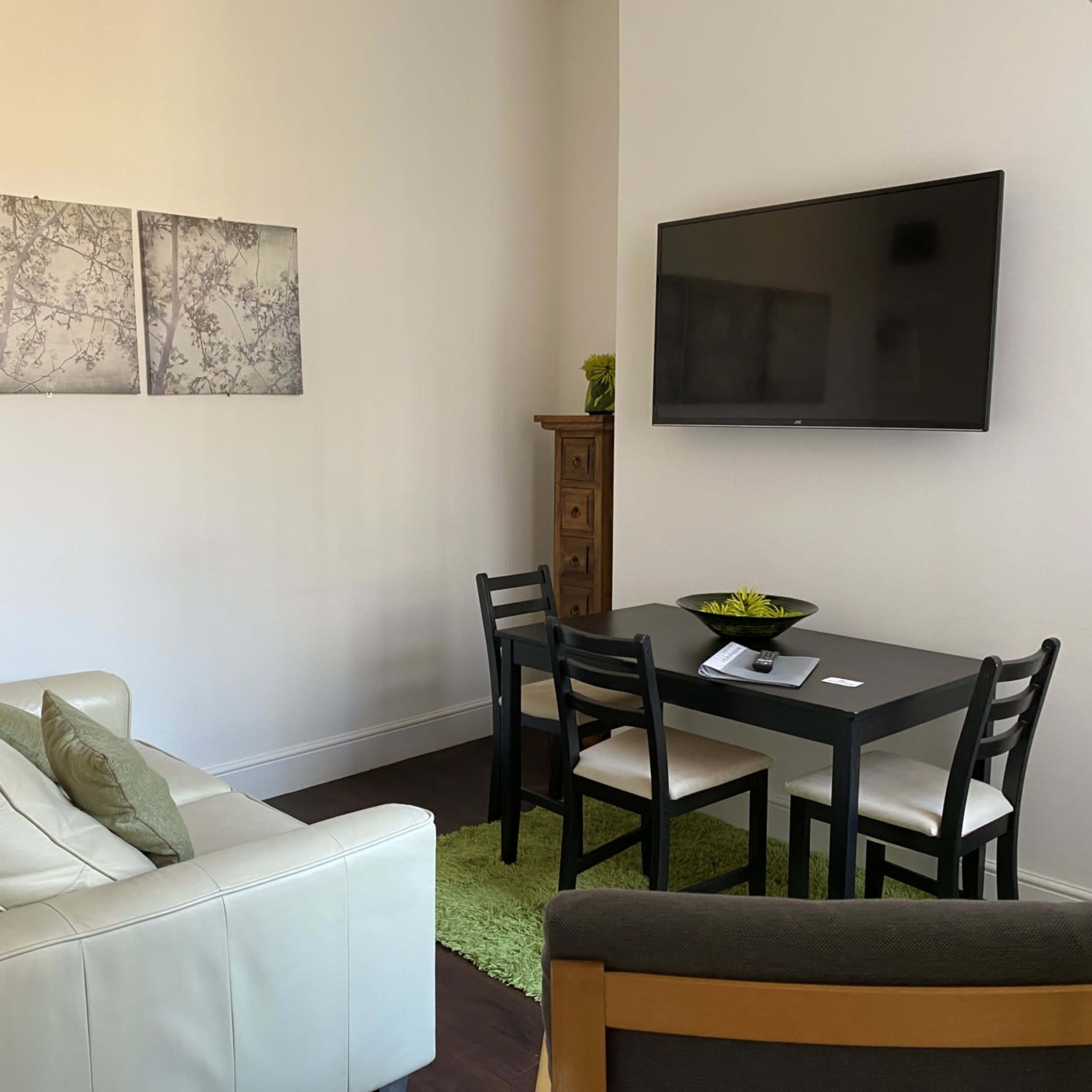 A small living area featuring a white couch, a black dining table with chairs, a television mounted on the wall, and three framed pictures.