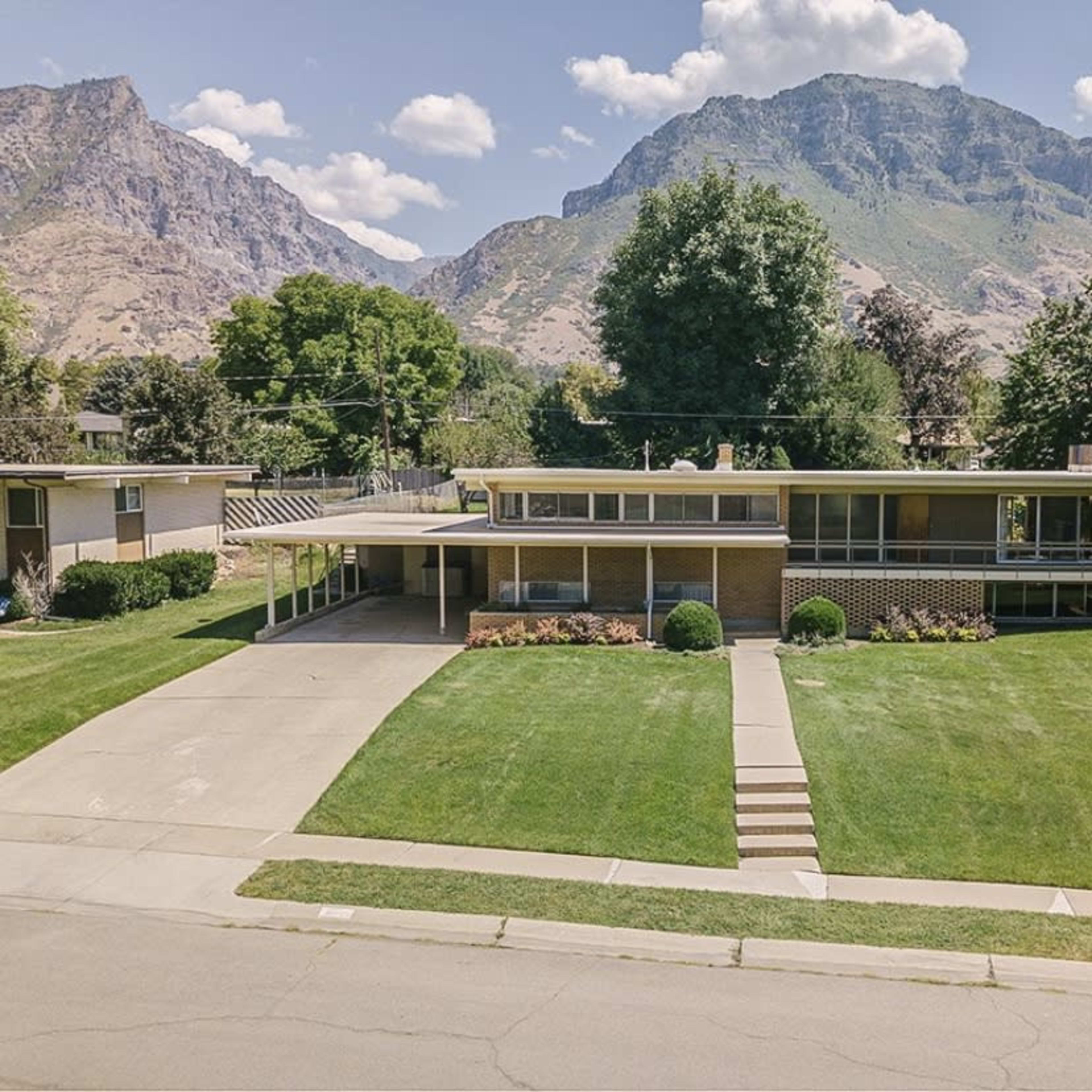 A modern, one-story house with a brick facade sits on a well-manicured lawn, framed by mountains in the background.