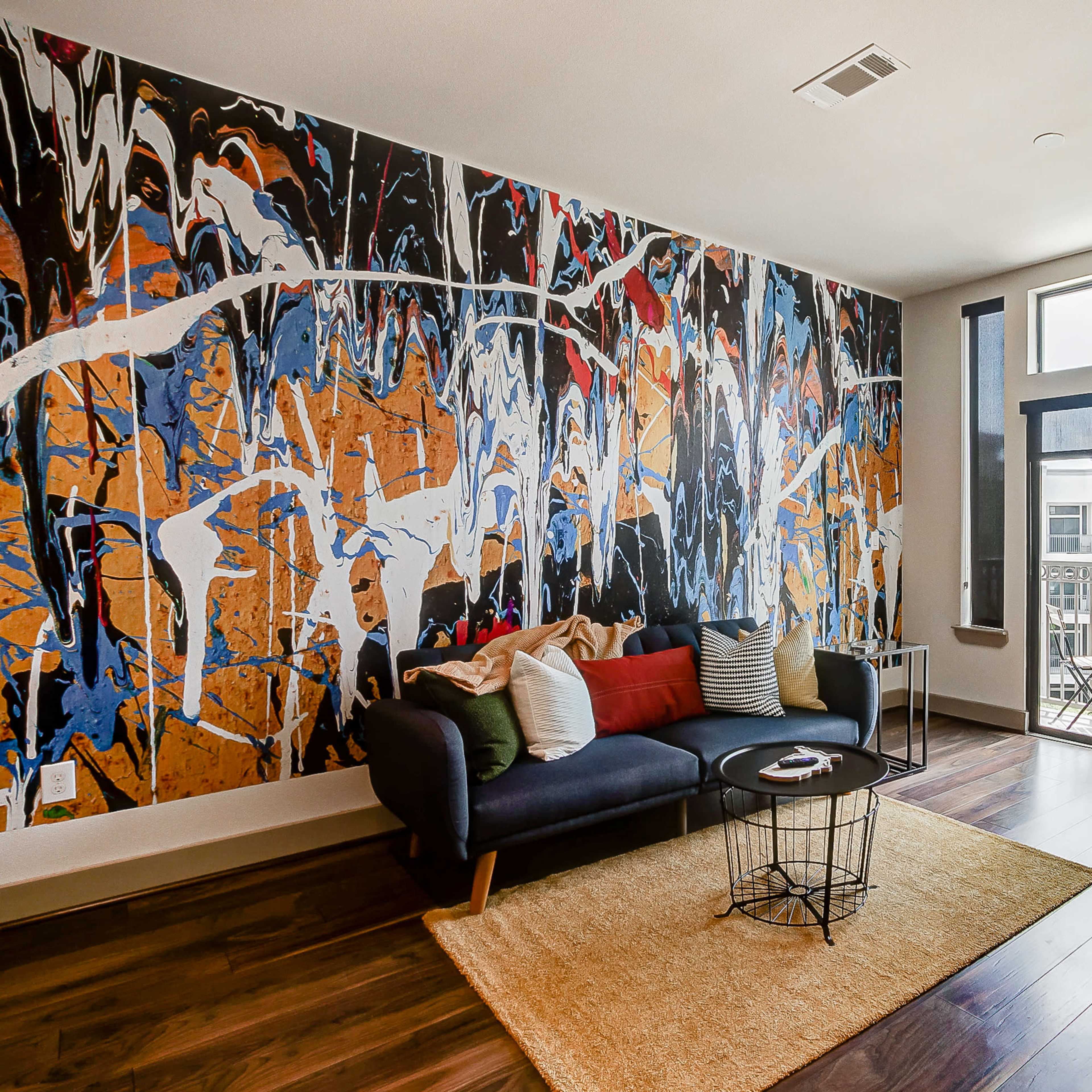 A living room with a large abstract mural on one wall, a dark sofa with colorful cushions, and a coffee table on a light rug, adjacent to a balcony visible through large windows.