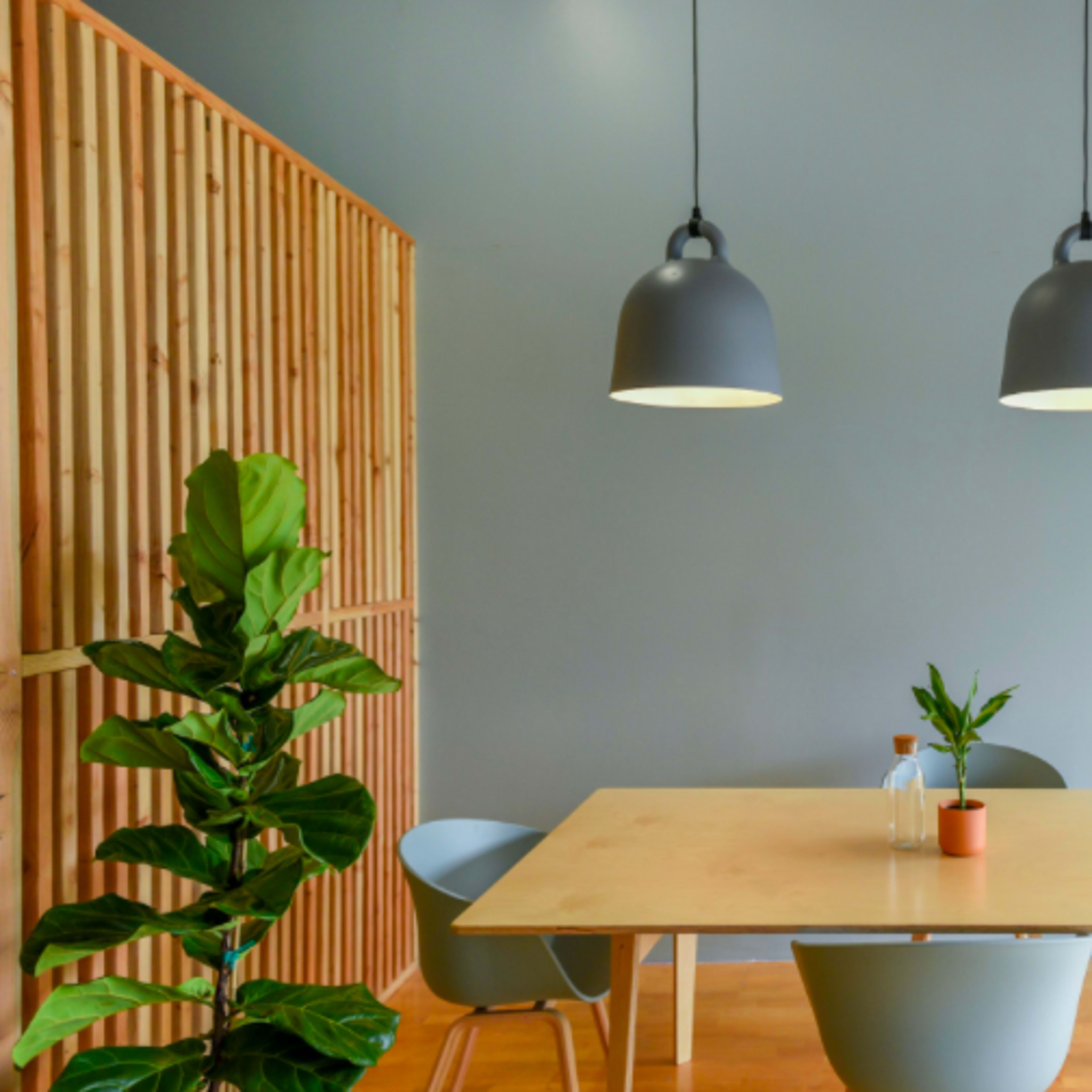 A modern dining area features a light wooden table surrounded by curved chairs, illuminated by two pendant lamps, with a potted plant in the corner.