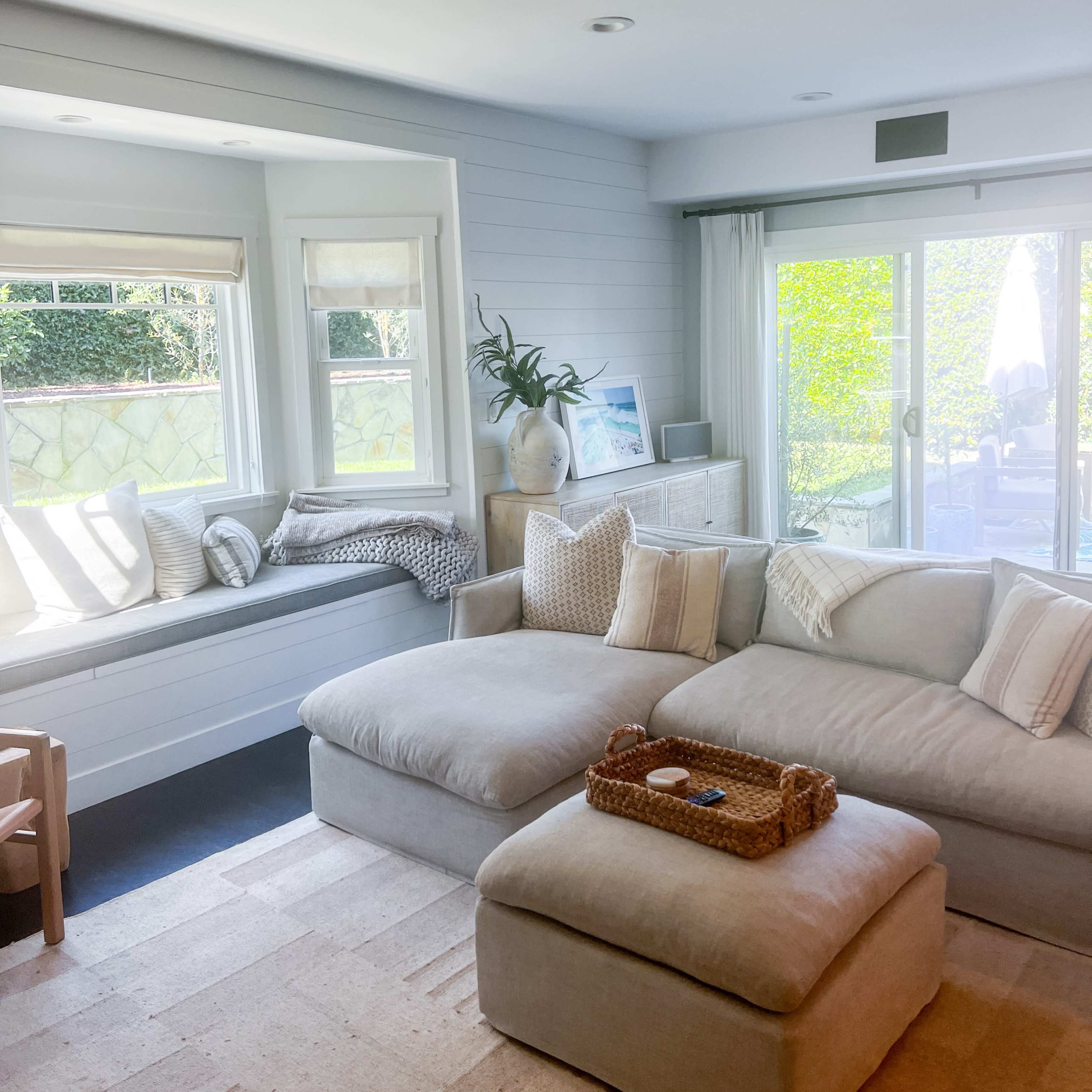A light-filled living room features a sectional sofa, a window seat, and a view of an outdoor patio through glass doors.
