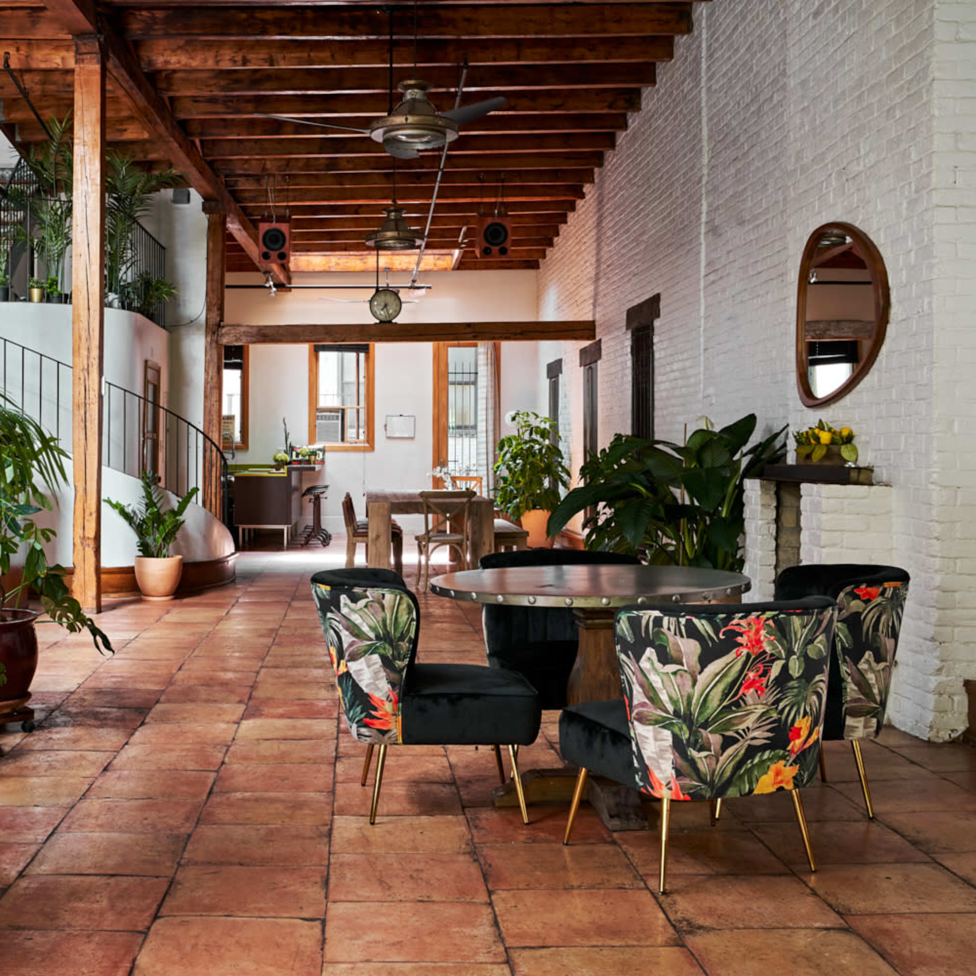 A spacious interior with terracotta tile flooring, a dining area featuring a round table and floral-patterned chairs, and large plants throughout the space.