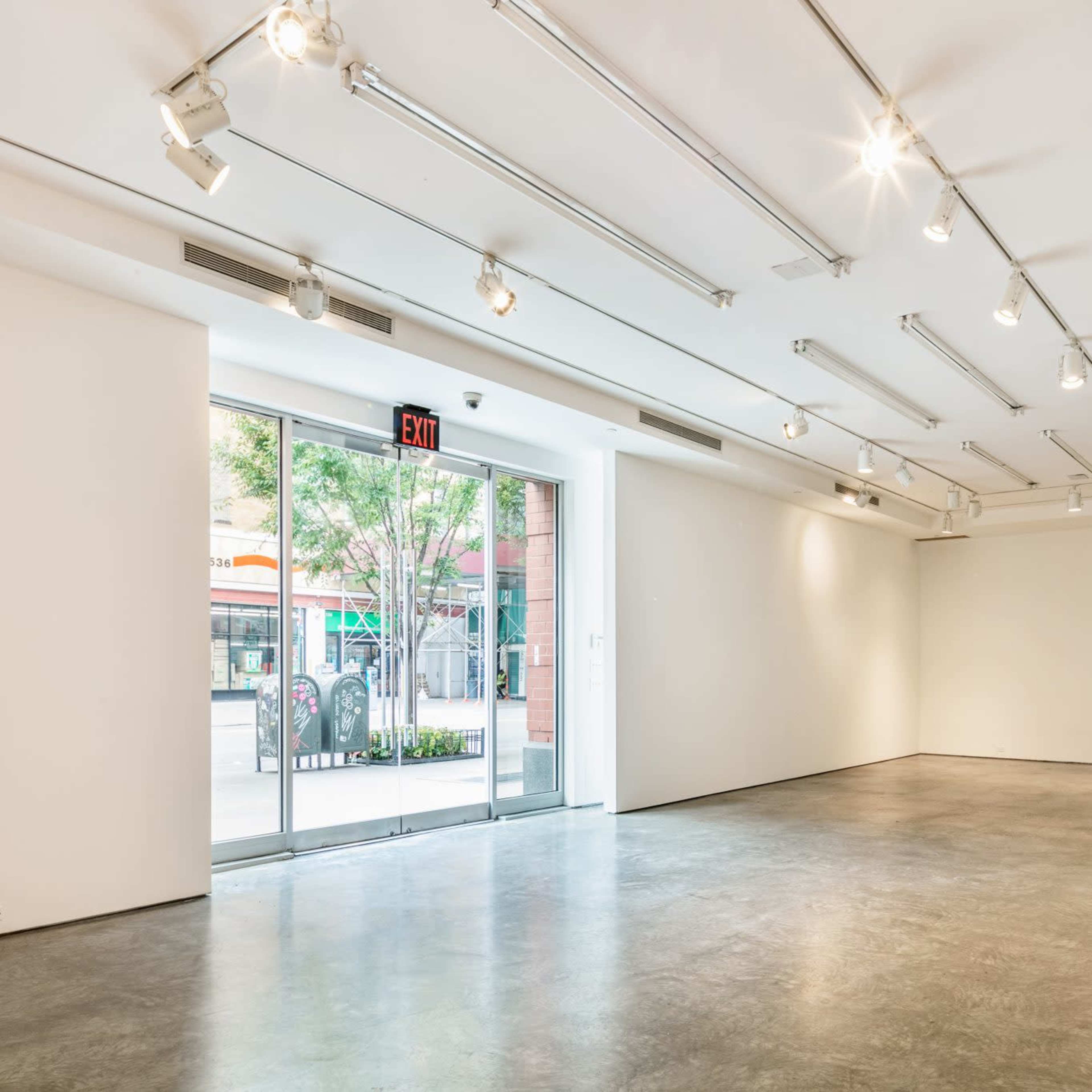 A spacious, well-lit gallery with white walls, concrete flooring, and large windows providing an exit to the outside.