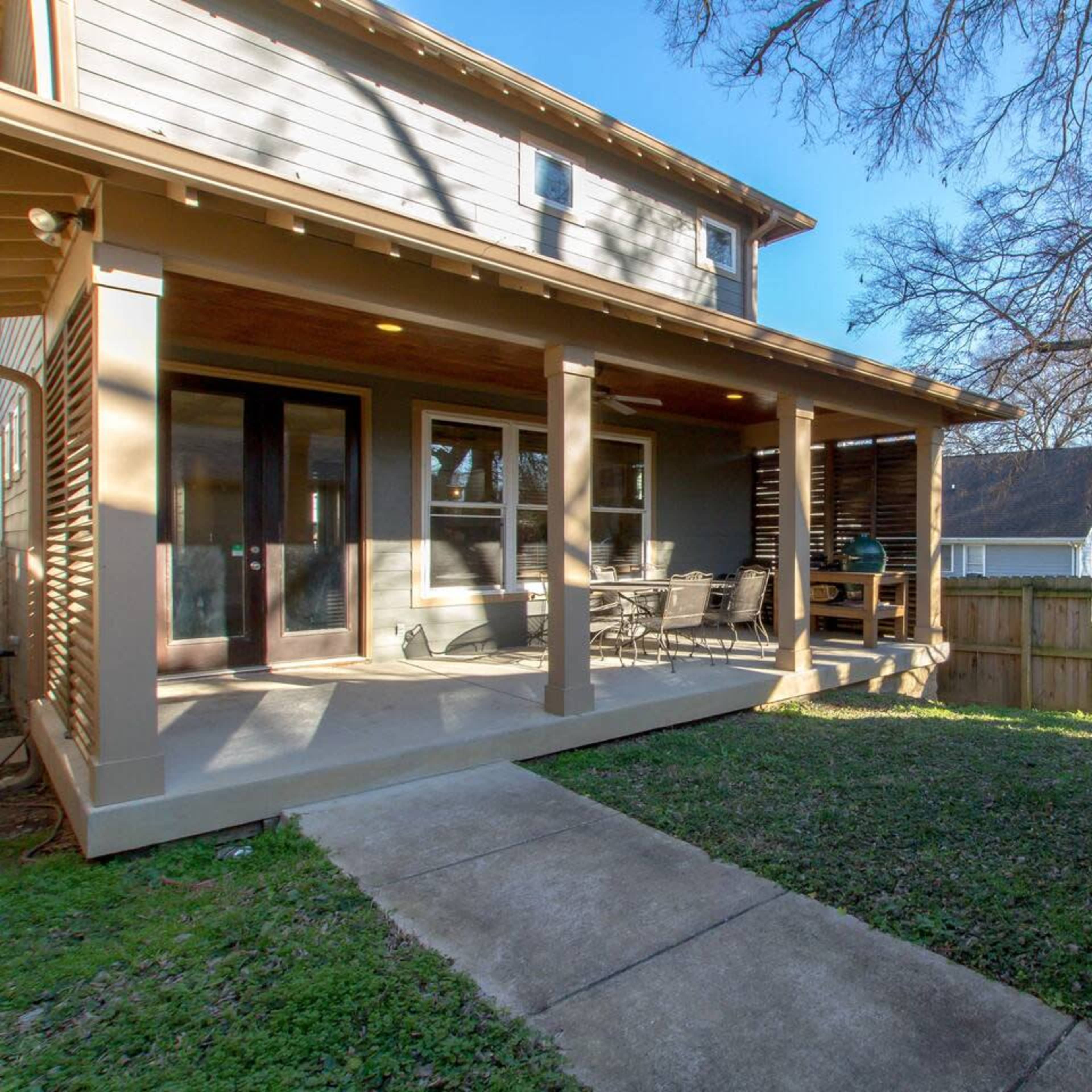 A backyard with a concrete pathway leading to a porch, surrounded by a wooden fence and several trees in the background.