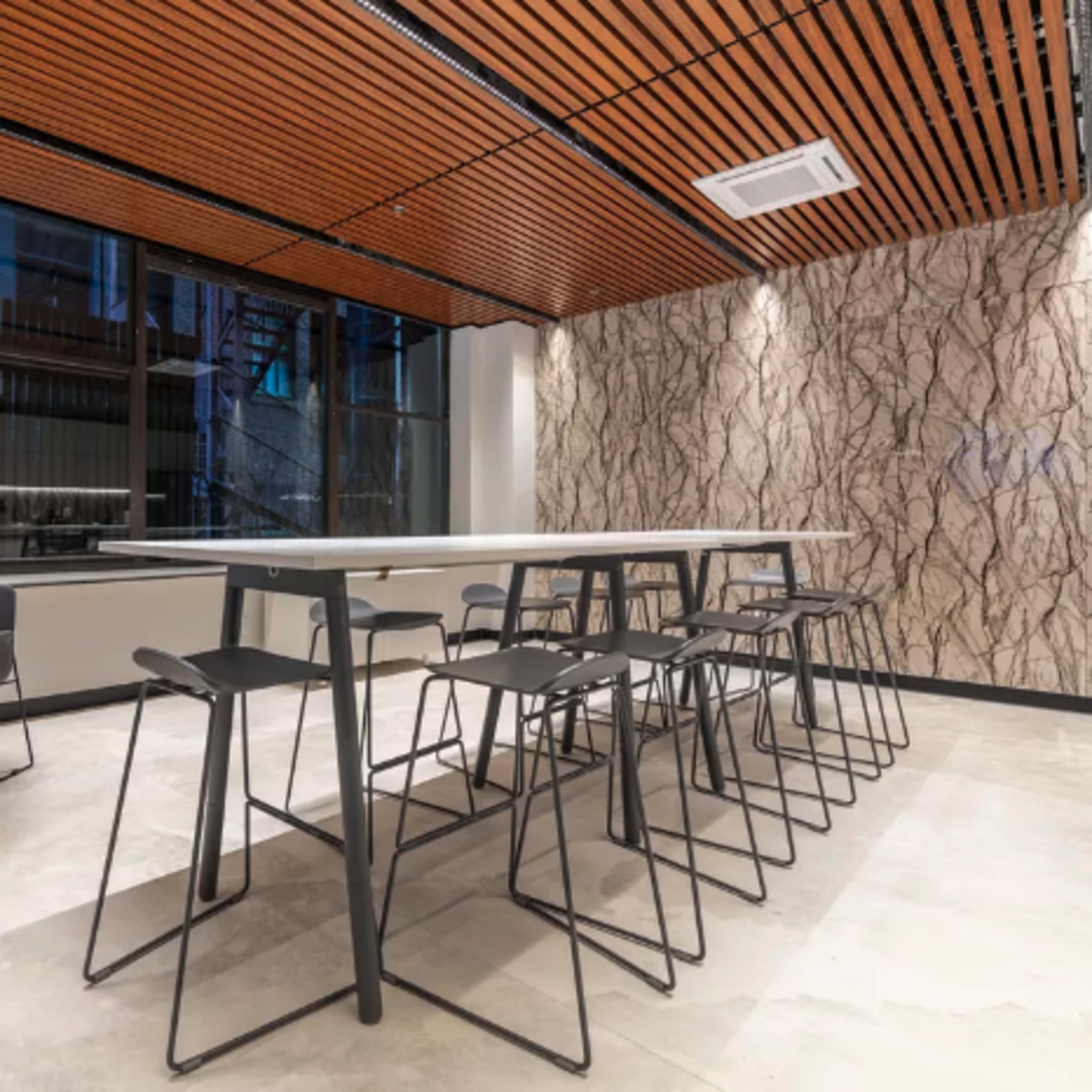 A modern dining area featuring long tables and black chairs, with a wood-paneled ceiling and textured wall.