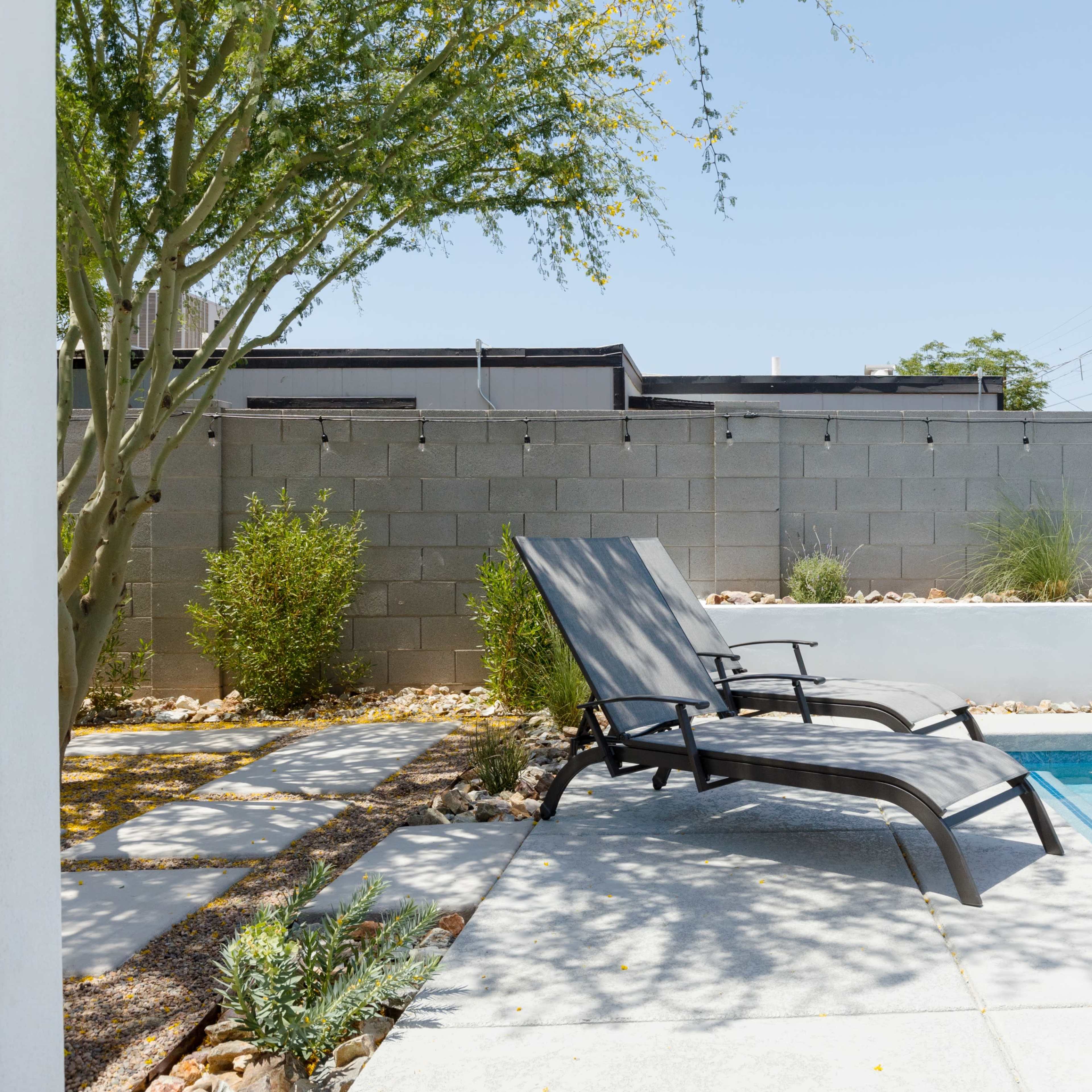 A pair of black lounge chairs sit beside a clear swimming pool, with a concrete wall and palm trees in the background.