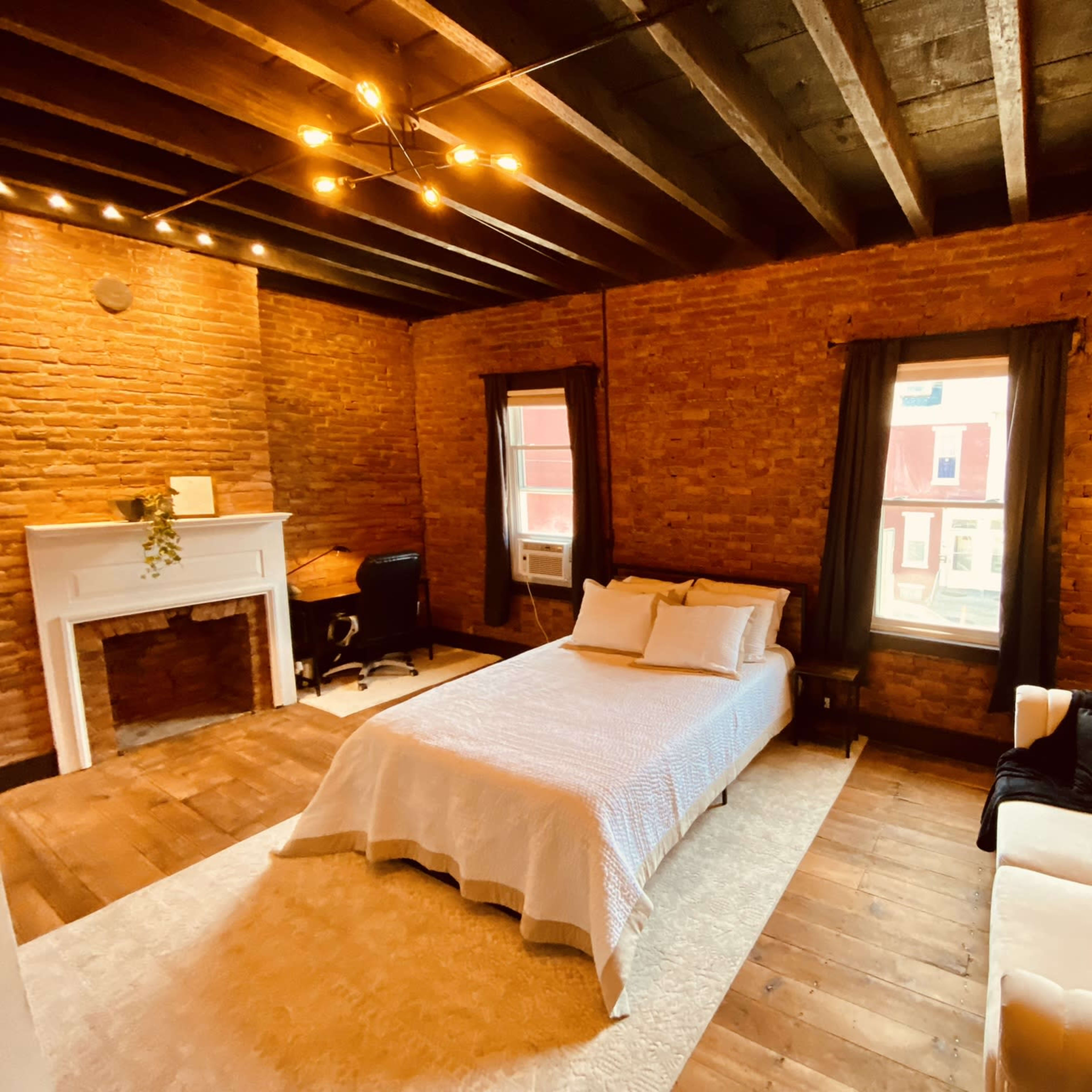 A rustic bedroom with exposed brick walls, a bed, a white sofa, a small desk, and large windows allowing natural light.