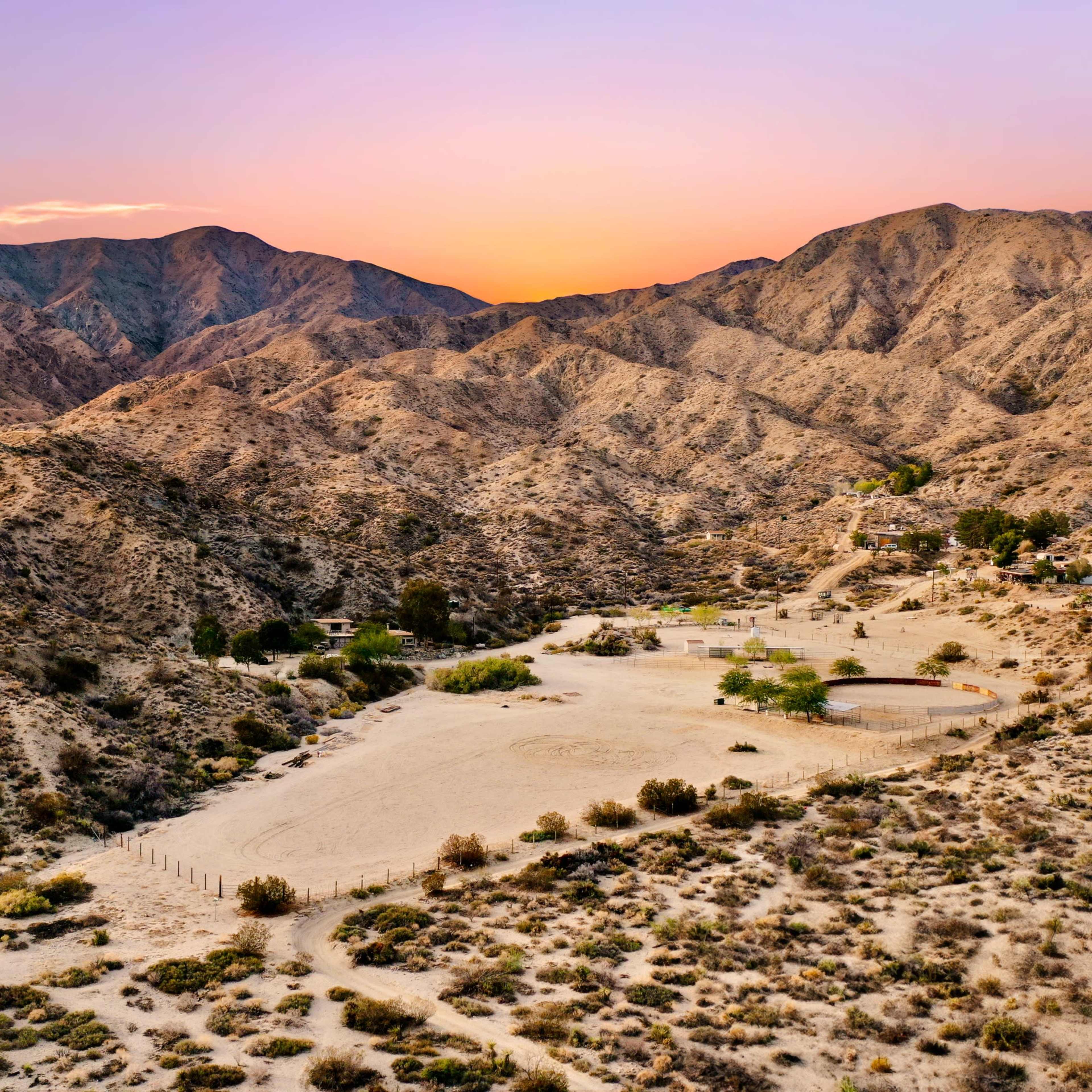 A desert landscape at sunset, featuring rugged mountains and a sparse settlement in the foreground.