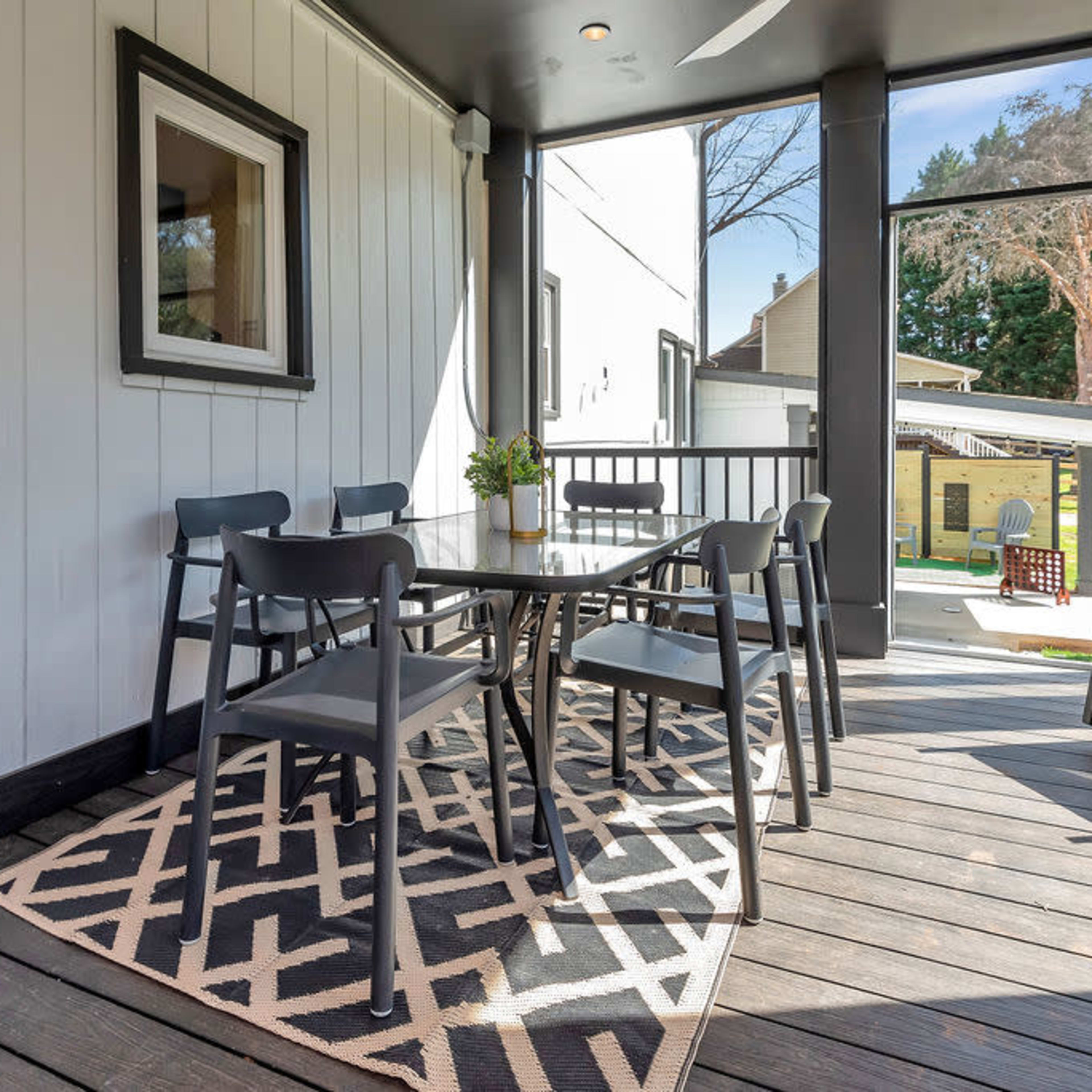 A patio area features a table and chairs on a patterned rug, with large glass windows and a view of a backyard.