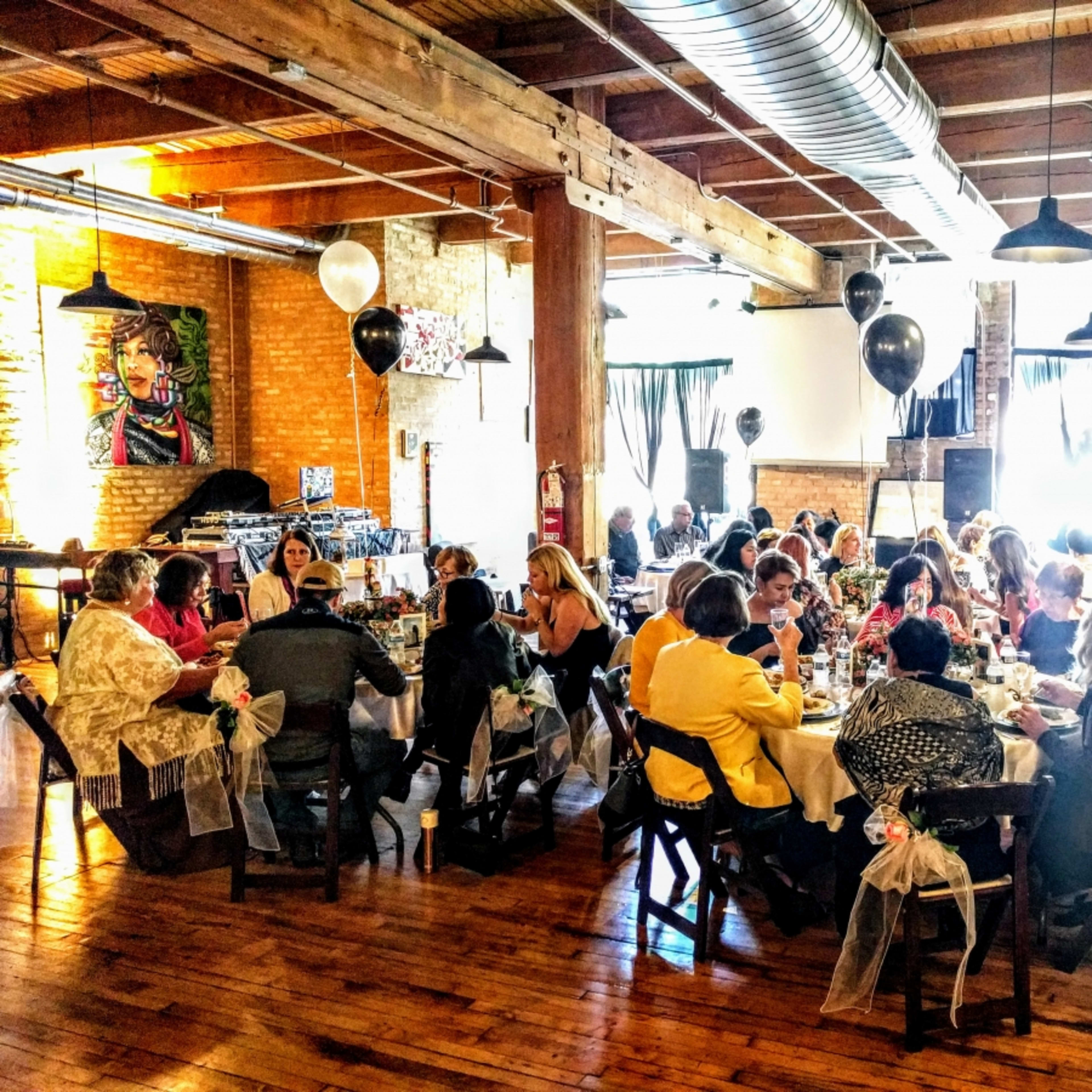 A group of women gathers around tables in a spacious, well-lit room decorated with artwork and balloons.