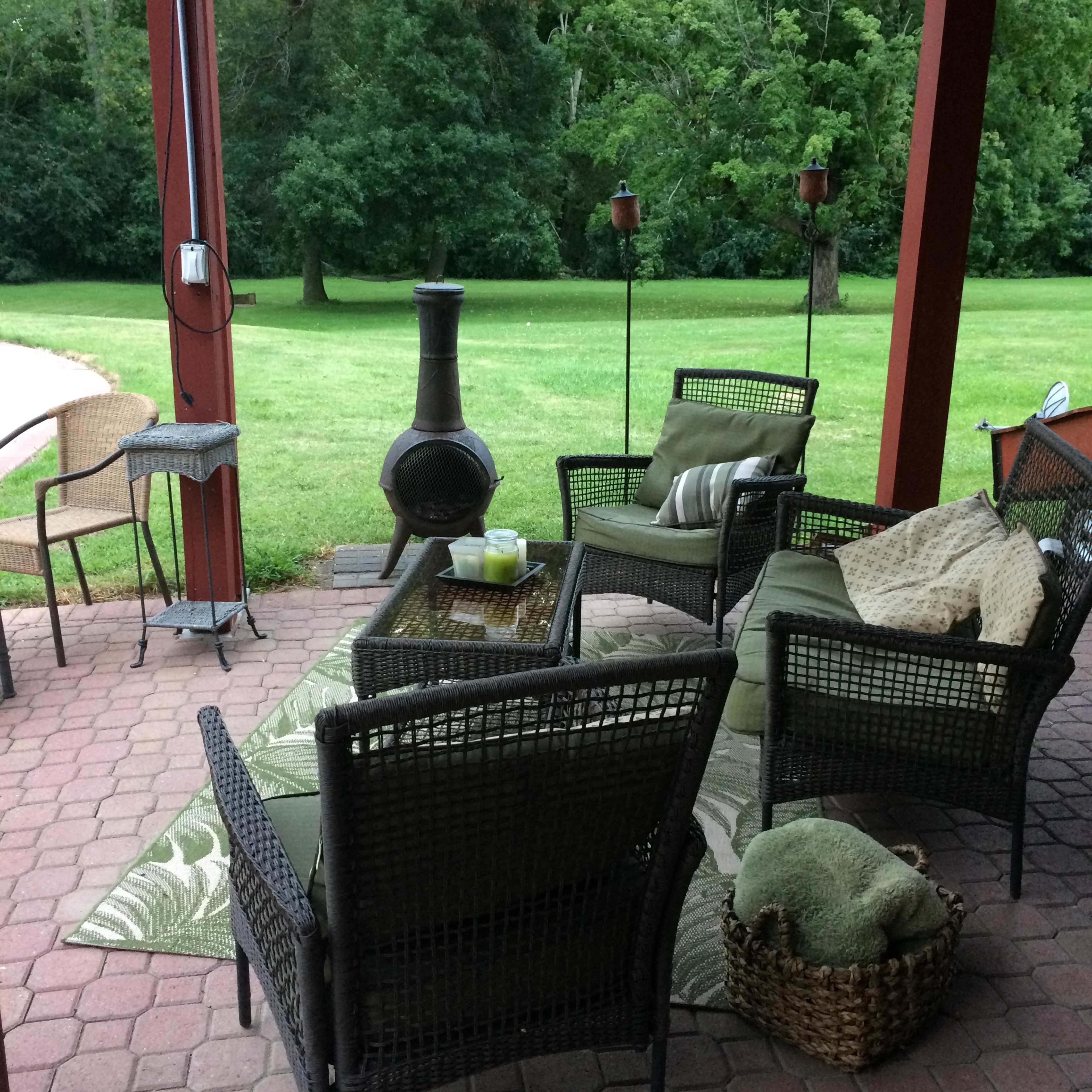 A cozy outdoor seating area features wicker chairs with cushions, a table, and a chiminea, set against a backdrop of lush greenery.