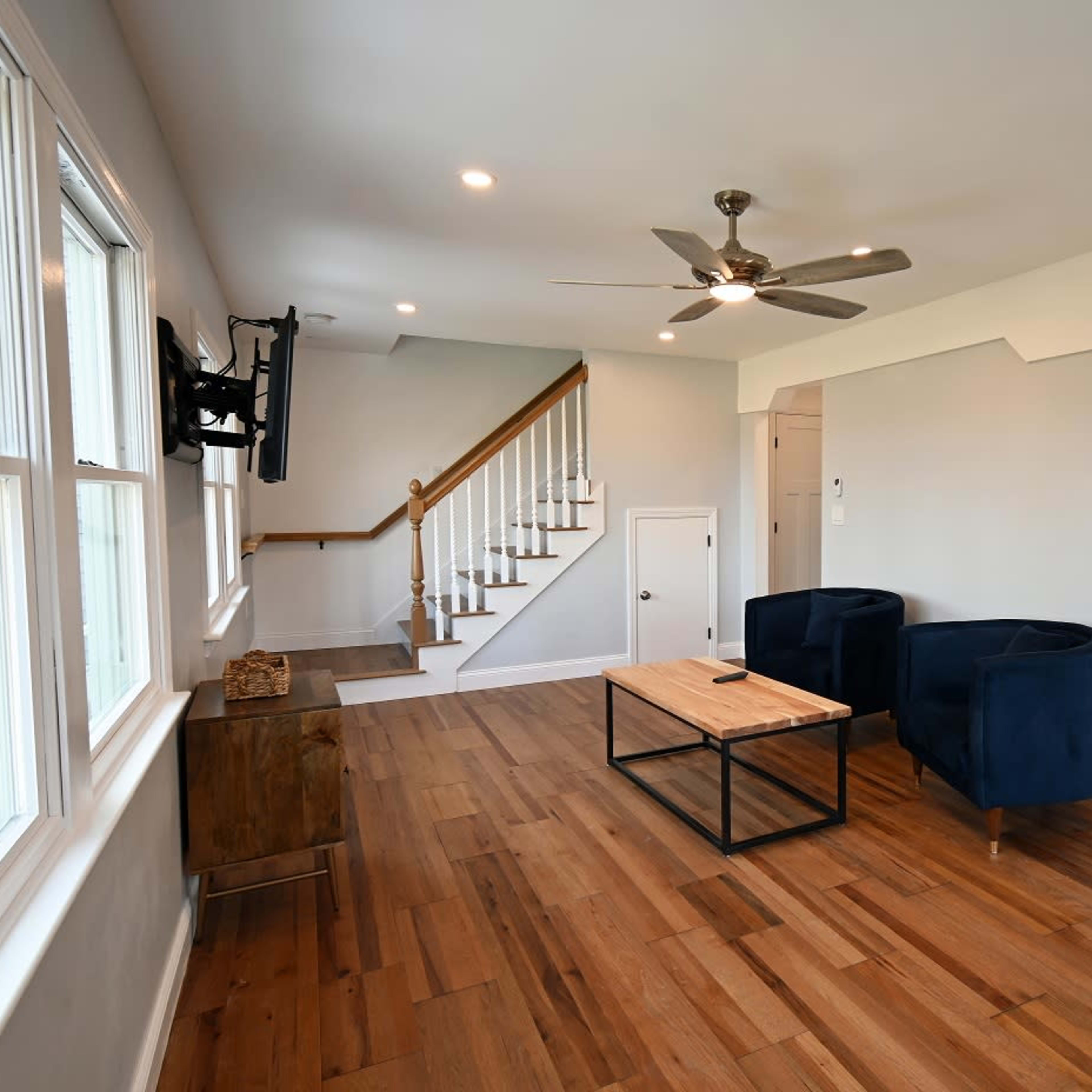 A modern living room features two blue chairs, a wooden coffee table, and a staircase leading to an upper level, with an open kitchen visible in the background.